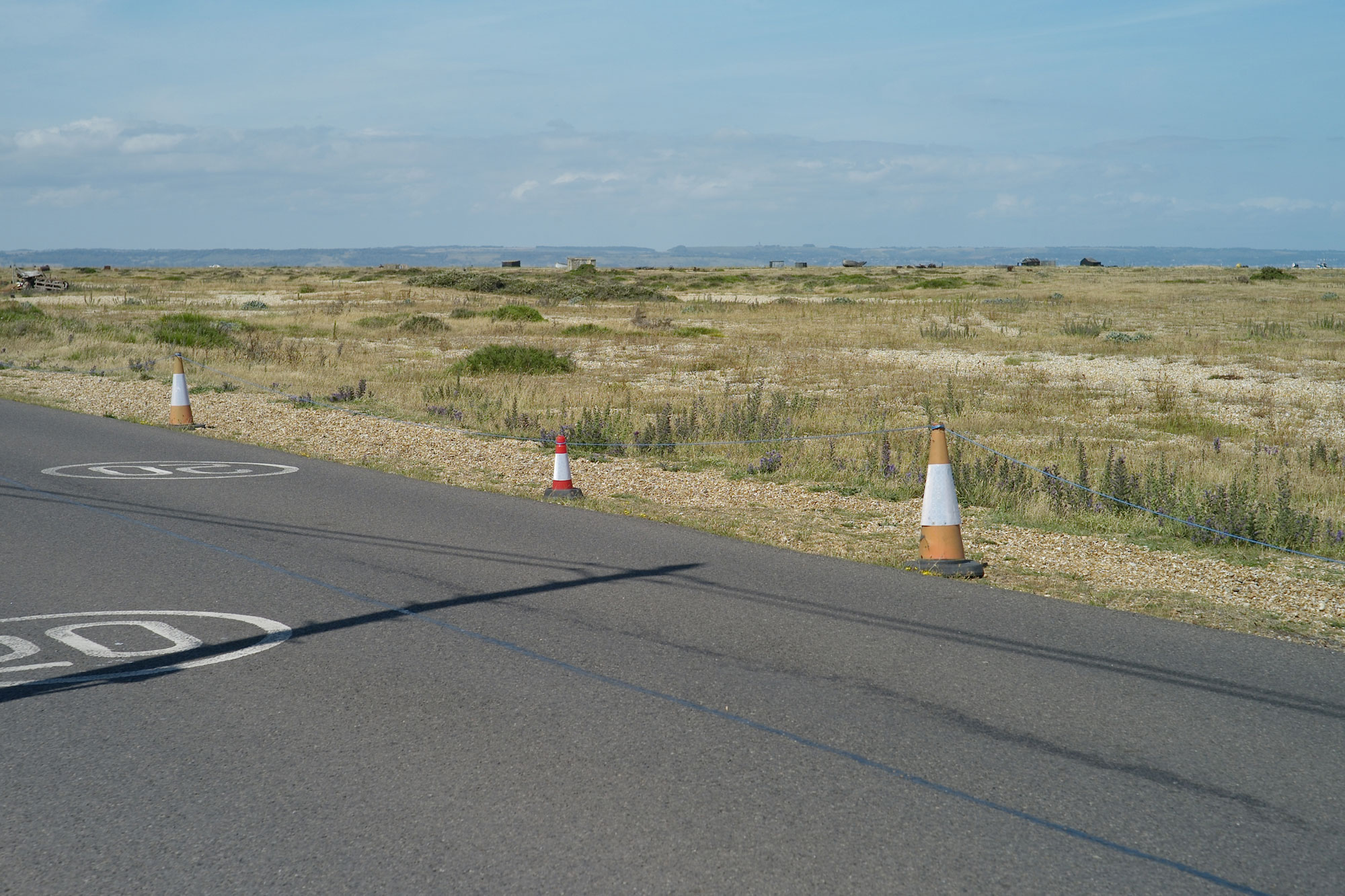 A road reading 20mph in Dungeness lined by traffic cones. Behind them, a desert-like landscape with foliage and cloudy blue skies. 