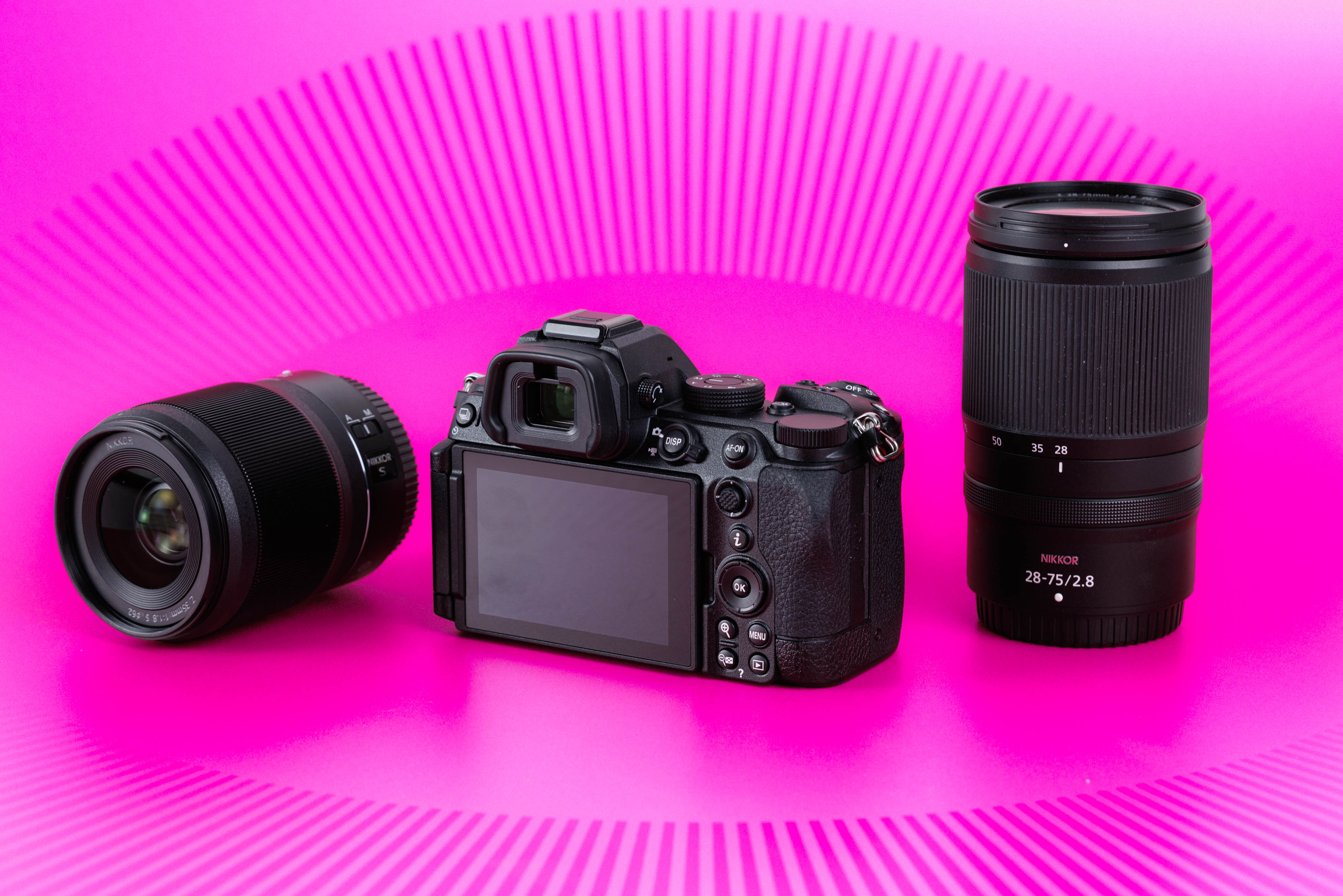 Studioaufnahme der Rückseite einer gebrauchten Nikon Z5 II zwischen zwei NIKKOR-Z-Objektiven vor leuchtend pinkem Hintergrund