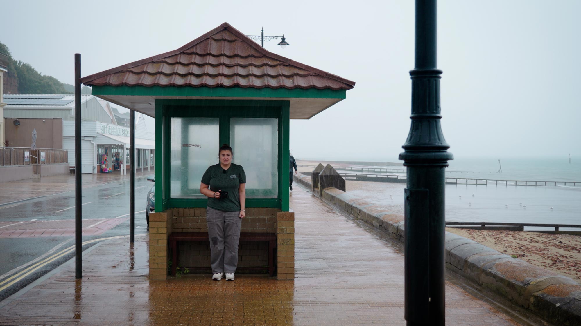 Amy Moore takes shelter, Leica Q3 in hand, on the seafront at the Isle of Wight. This is a screenshot from the review on the MPB YouTube channel.