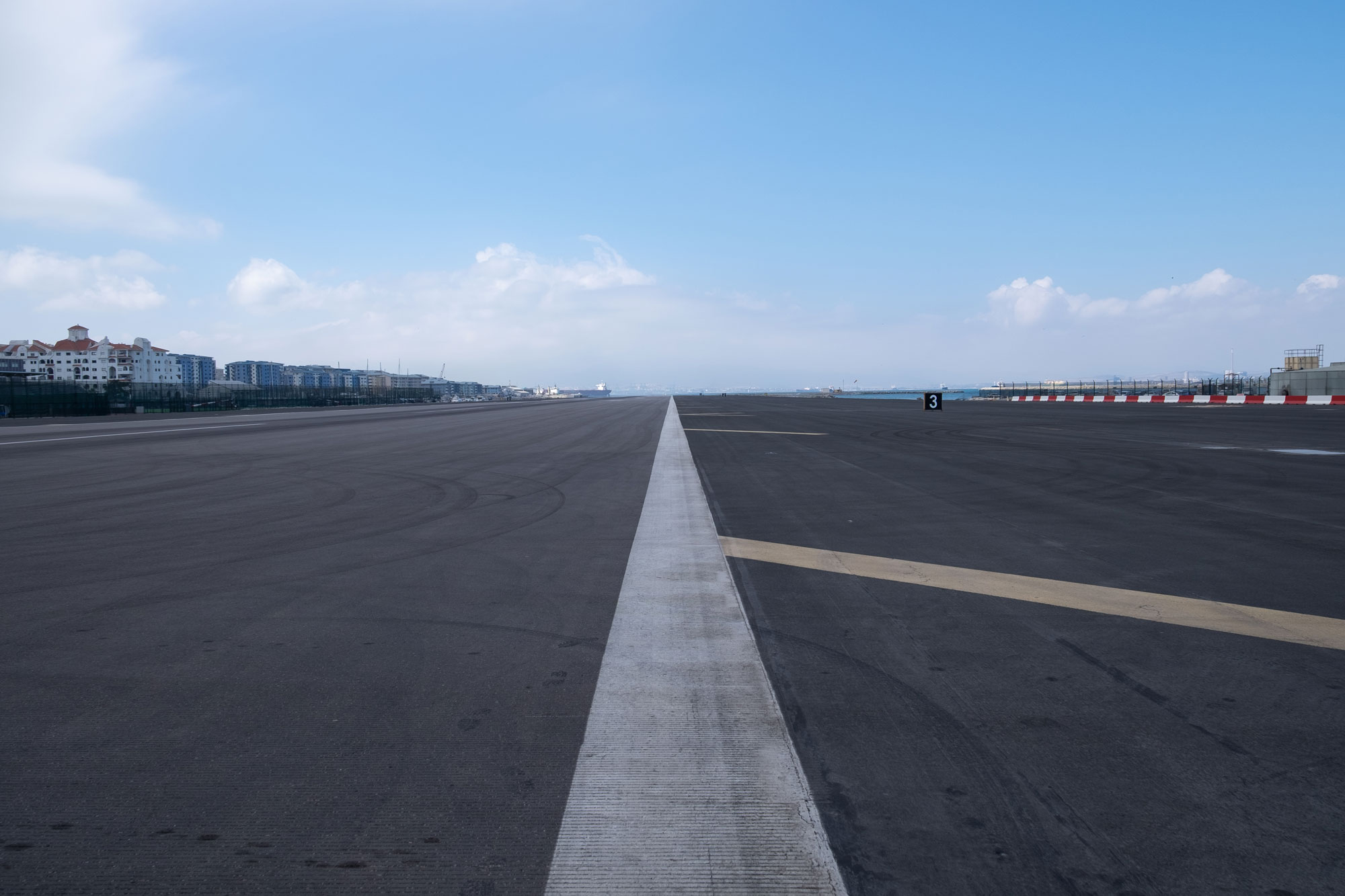 A long, straight white line stretches into the distance on an empty runway for aeroplanes. A blue sky with white clouds fills the background.
