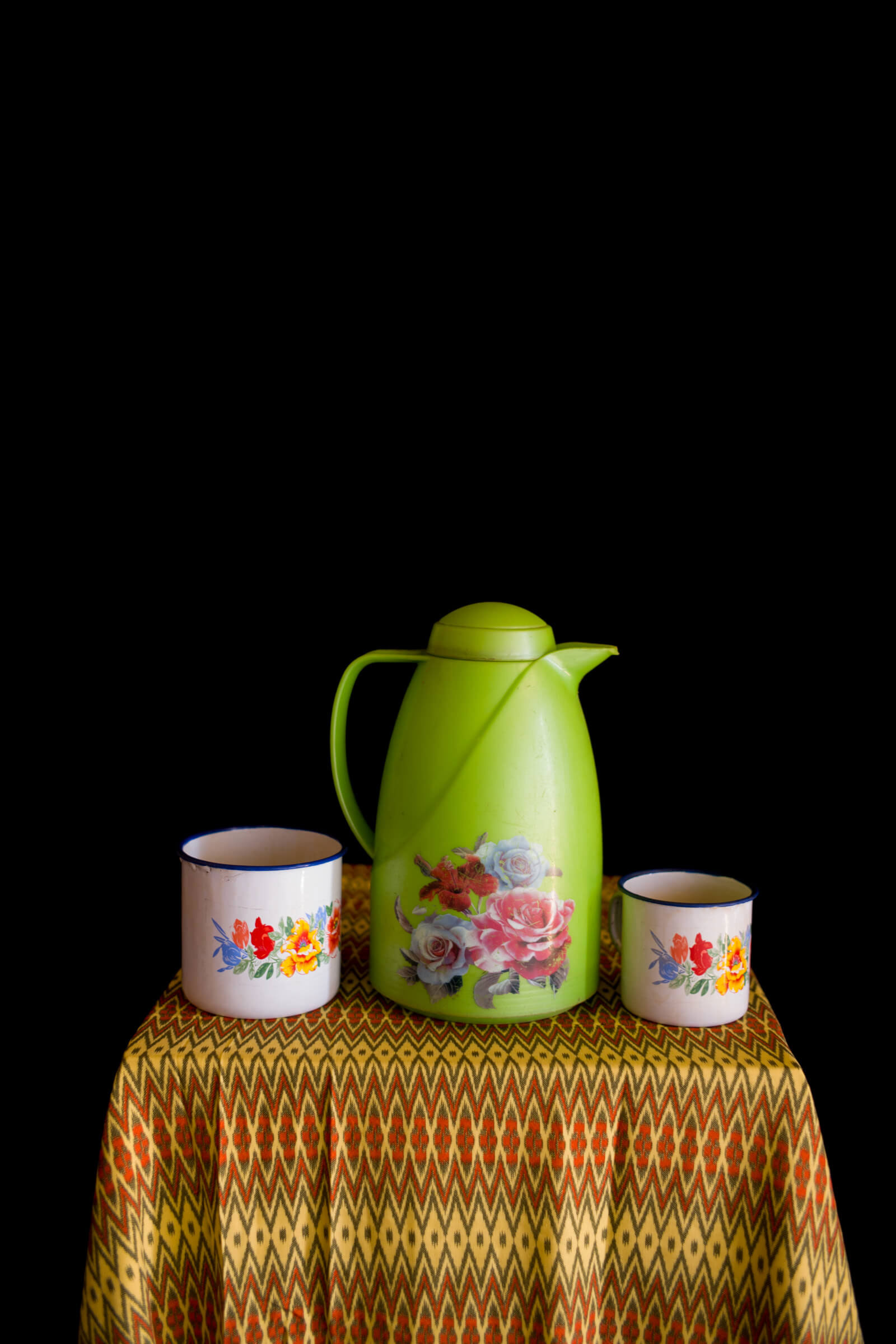 Ein passendes Set von Tassen und einer Kaffeekanne mit Blumenmuster stehen auf einer bunten Tischdecke vor einem schwarzen Hintergrund.