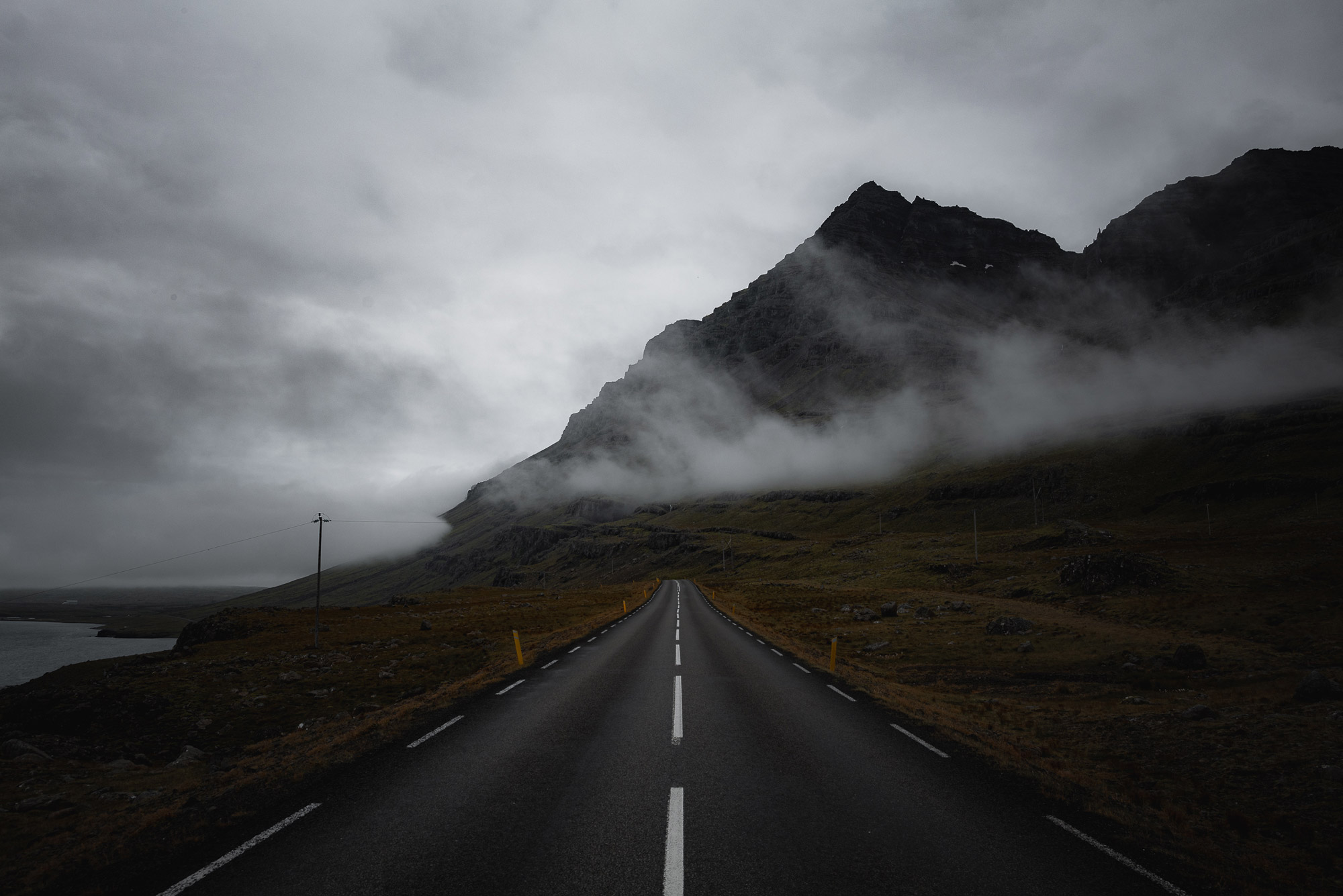 A road with white lines in the middle stretching into the distance, with mountains and clouds in the background, shot by Paul Watson with a Panasonic S1R