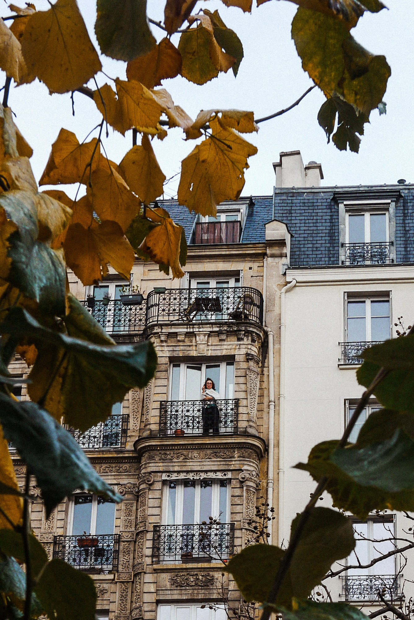 Streetaufnahme einer Person, die auf einem Balkon mit schmiedeeisernem Geländer eines Pariser Wohnhauses steht. Im Vordergrund herbstliche Blätter. Aufgenommen von Wesley Verhoeve mit einer Panasonic Lumix GF1.