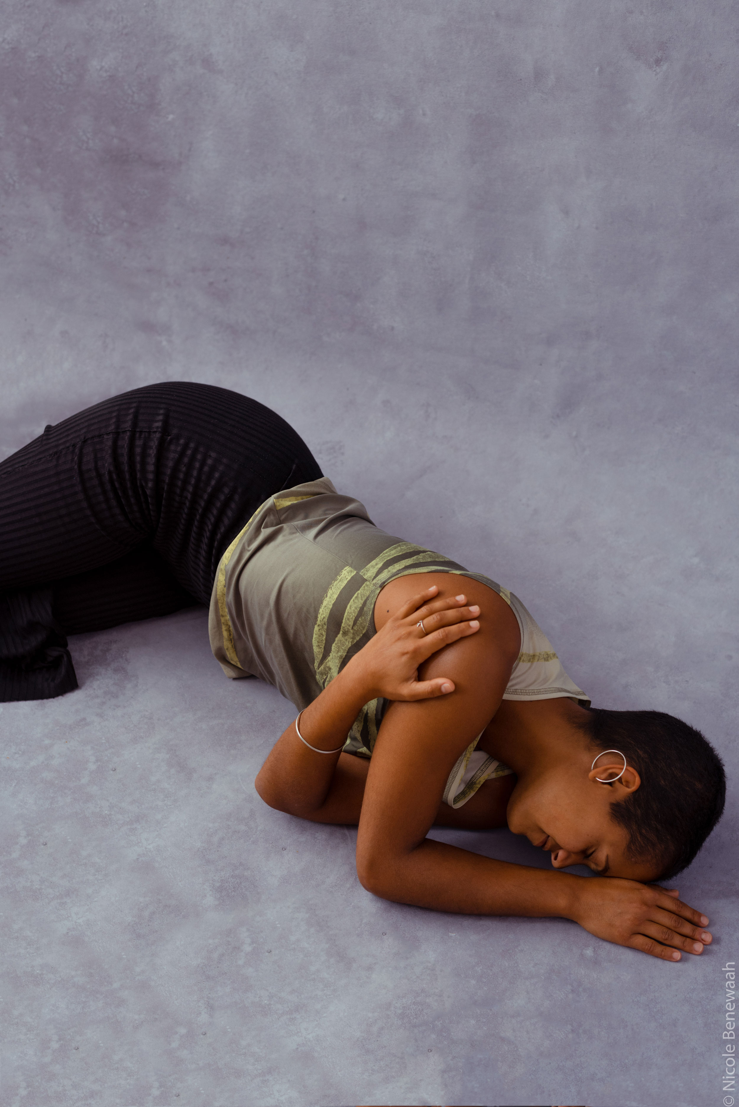 Femme noire allongée sur un sol violet et se serrant contre elle avec son bras gauche, photographiée par Nicole Benewaah