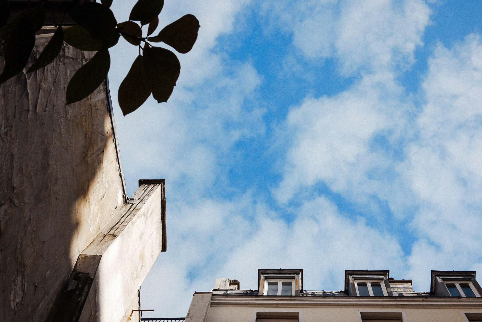 An upward street photograph showing the edges of Paris building facades and rooftop windows framed against a bright blue sky with scattered clouds, shot by Wesley Verhoeve.