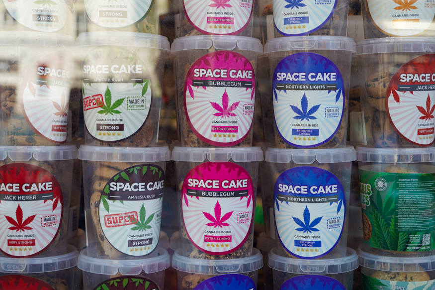 Een kleurrijke uitstalling van spacecakes in een Amsterdamse etalage, met opvallende labels en stapels bakken vol iconische cannabisbranding, gefotografeerd door Louise Kluit