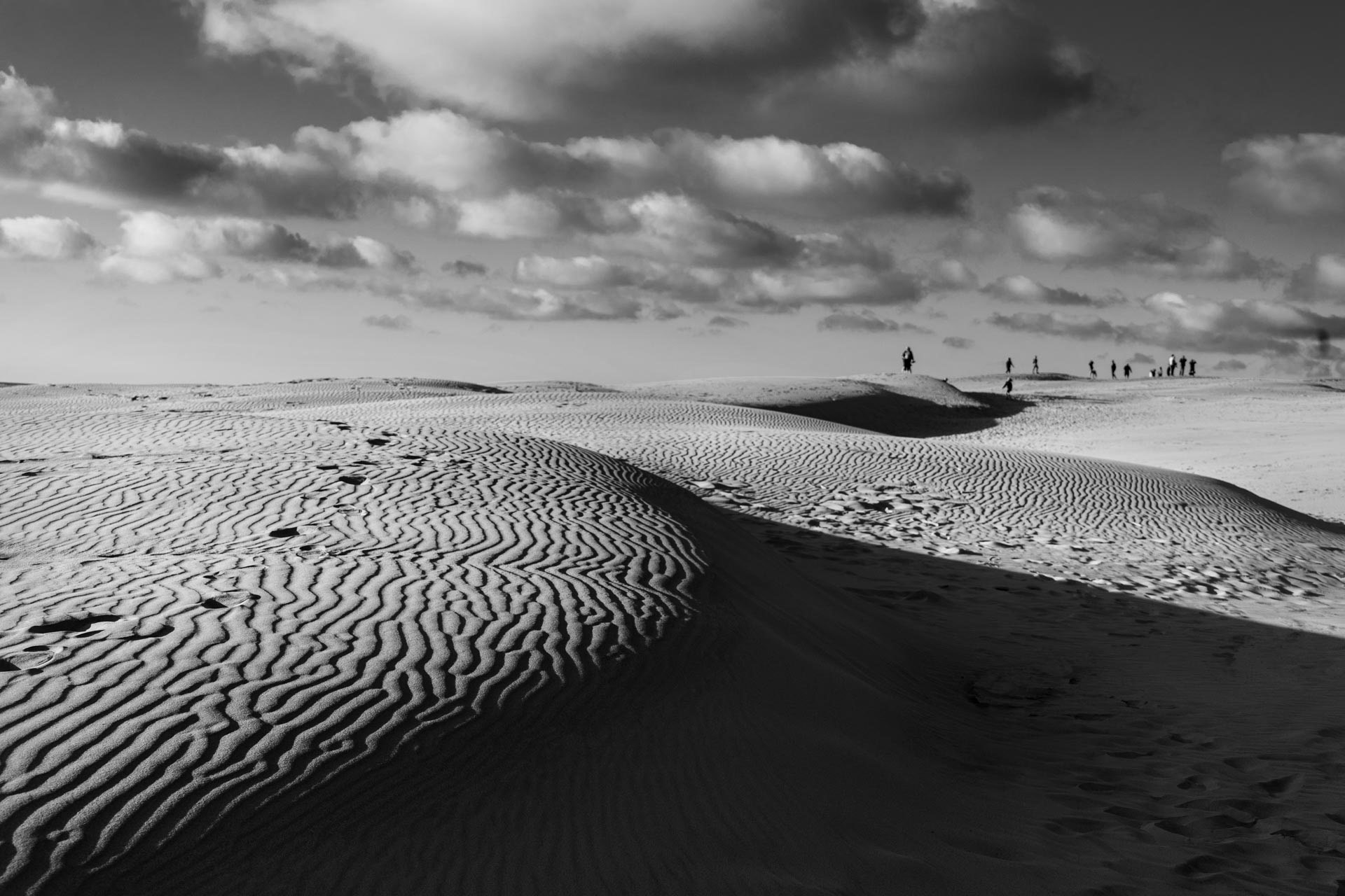 Zwart-witfoto van het migrerende duin Råbjerg Mile in Denemarken met wandelende figuren aan de horizon onder een bewolkte lucht, gefotografeerd door Maike Jarsetz.