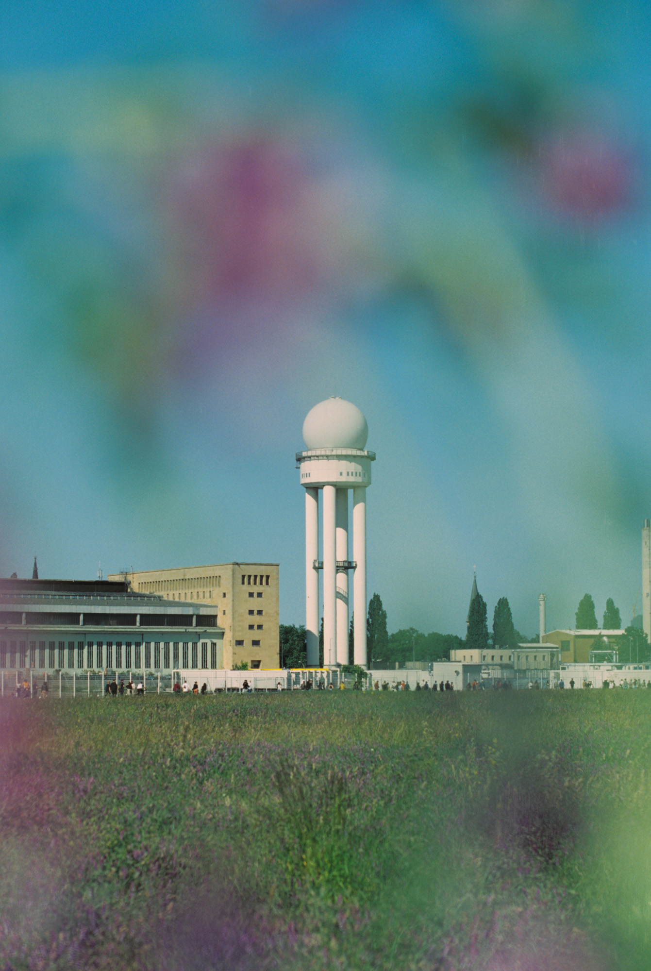 Dreamy film photograph of the white radar tower rising above Berlin’s former airport at Tempelhofer Feld, framed by soft-focus wildflowers and summer haze. Shot by Austin Fassino on 35mm film.