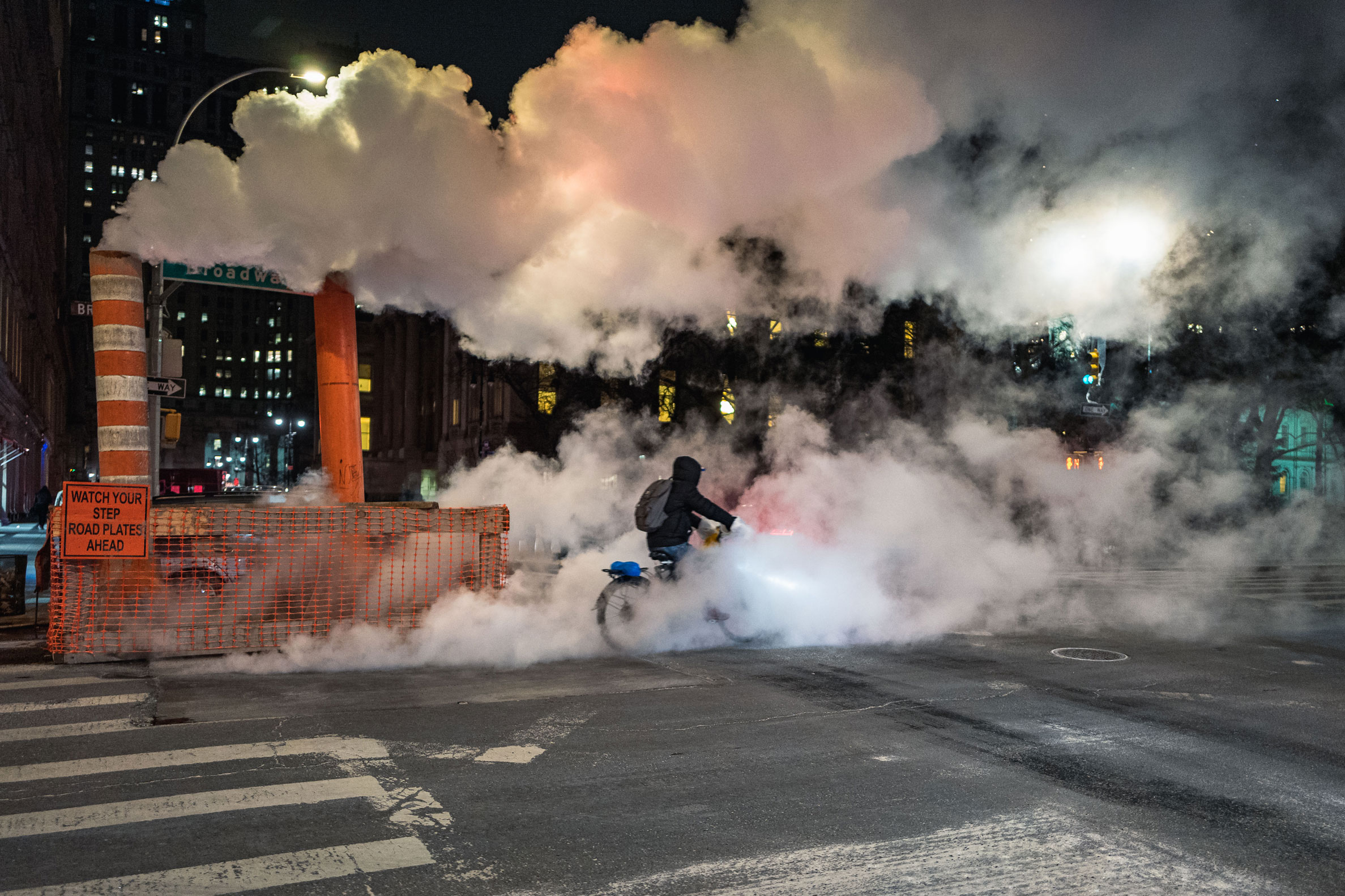Une personne circule à vélo dans une rue urbaine la nuit avec de la fumée. Photo prise par Manuel Pena avec un Leica Q.

