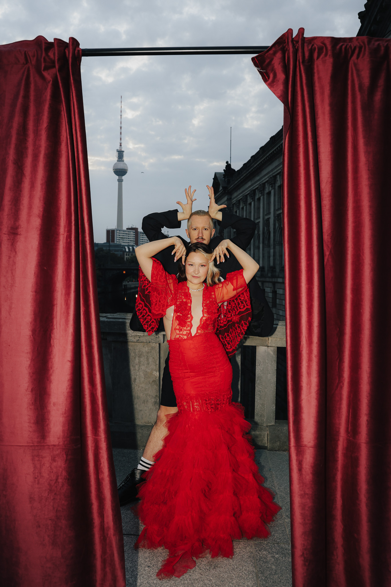 Creative portrait of a couple posing in vibrant red couture, blending art, fashion, and theatrical flair. In the background: Berlin’s Museumsinsel and iconic TV tower. Shot by Marina Polovinkina on a Canon EOS R6 Mark II.