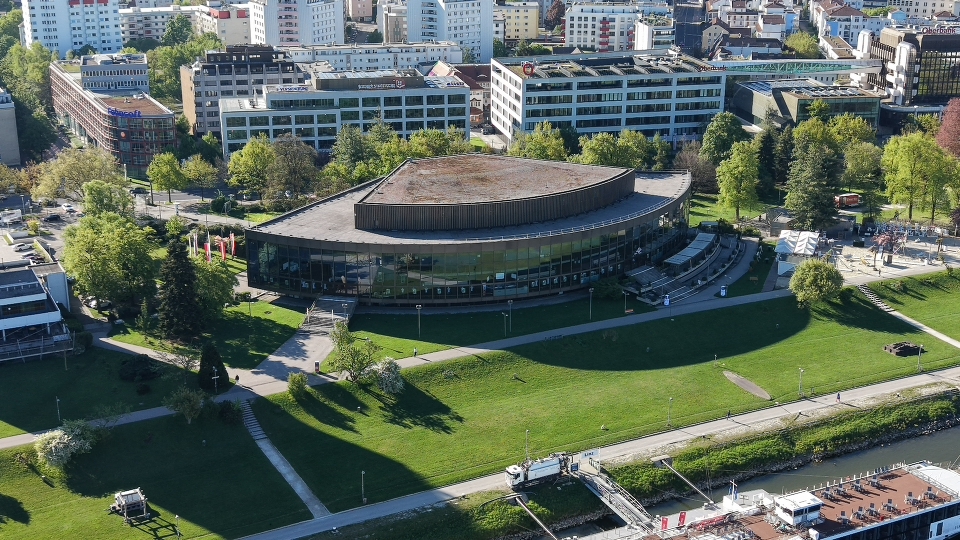 Imagem aérea captada com o DJI Mini 5 Pro, mostrando um edifício urbano rodeado por zonas verdes, ruas e edifícios na margem do rio. A composição destaca a escala da cidade e a capacidade do drone para captar enquadramentos amplos em contexto urbano.