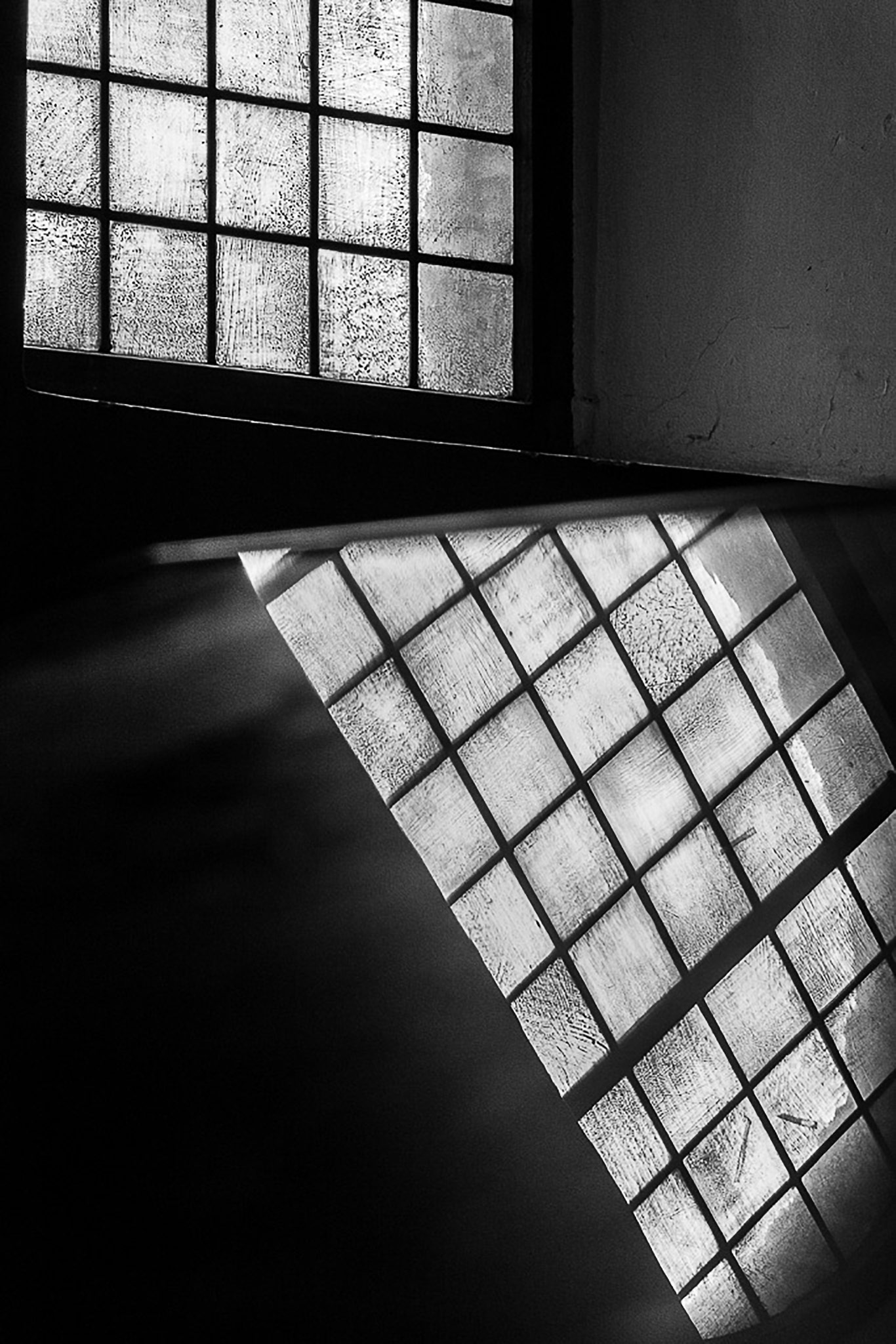 Schwarz-Weiß-Foto von Licht, das durch ein vergittertes Fenster scheint, das sich in einem Spiegel am unteren Rand des Bildes spiegelt, Foto von Maike Jarsetz