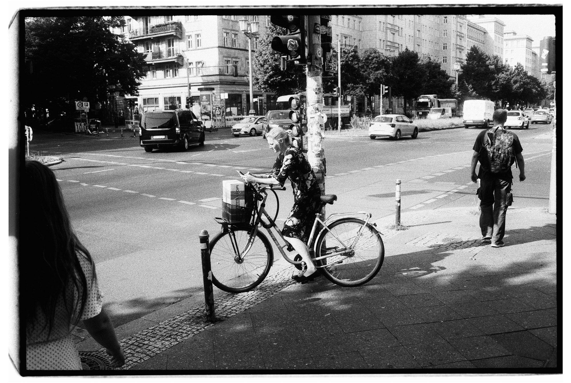 Black-and-white street photography of a cyclist waiting at a busy crossing on Berlin’s Karl-Marx-Allee, framed by pedestrians, traffic and high-contrast urban light. Shot by Verhoeve on 35mm film.