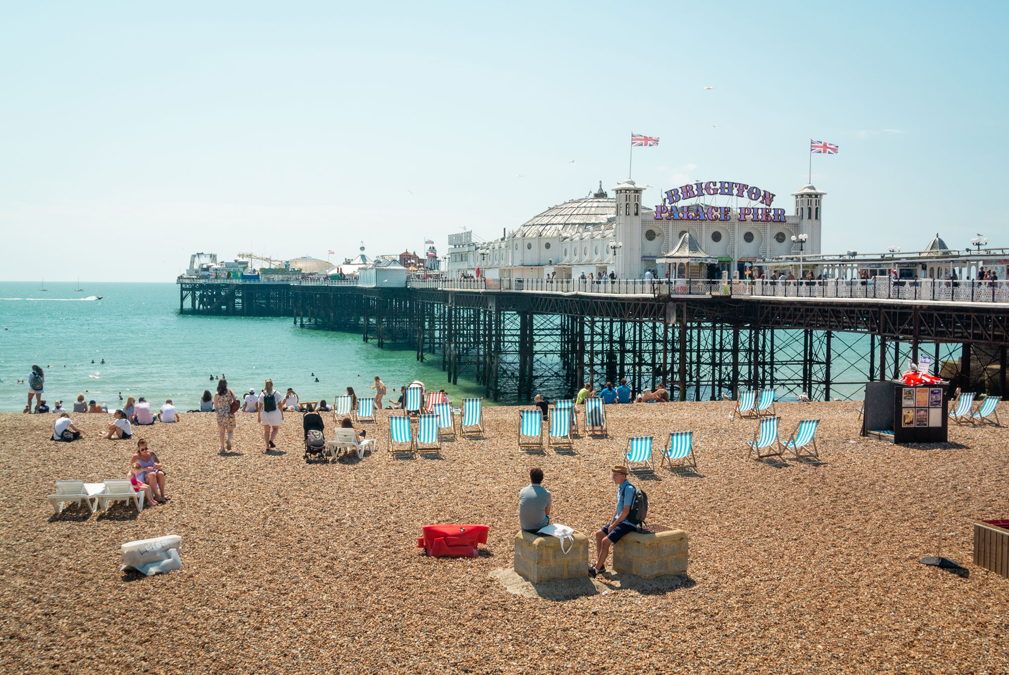 Ein gut besuchter Kiesstrand an einem Sommertag, mit dem Brighton Palace Pier im Hintergrund. Mehrere Menschen sonnen sich, während leere Liegestühle über den Strand verteilt sind.