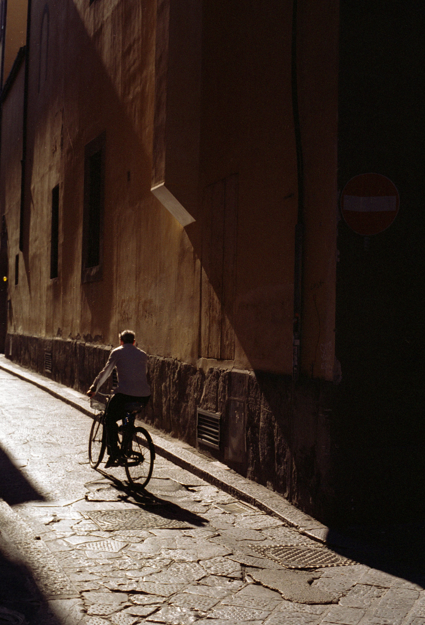 A person riding a bicycle down a small narrow street with harsh light filling the space by Ian Howorth