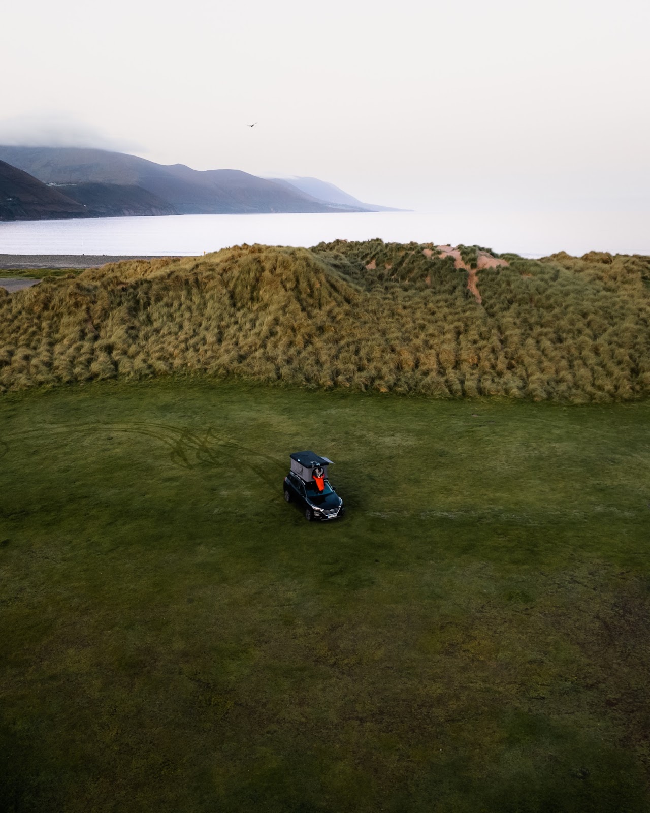 A drone photo of a person, wearing a red coat, standing on the roof of their car, which is parked in a green field surrounded by trees, mountains and the sea in the background, shot by Jonas Hanspach with a DJI Mavic Air 2S