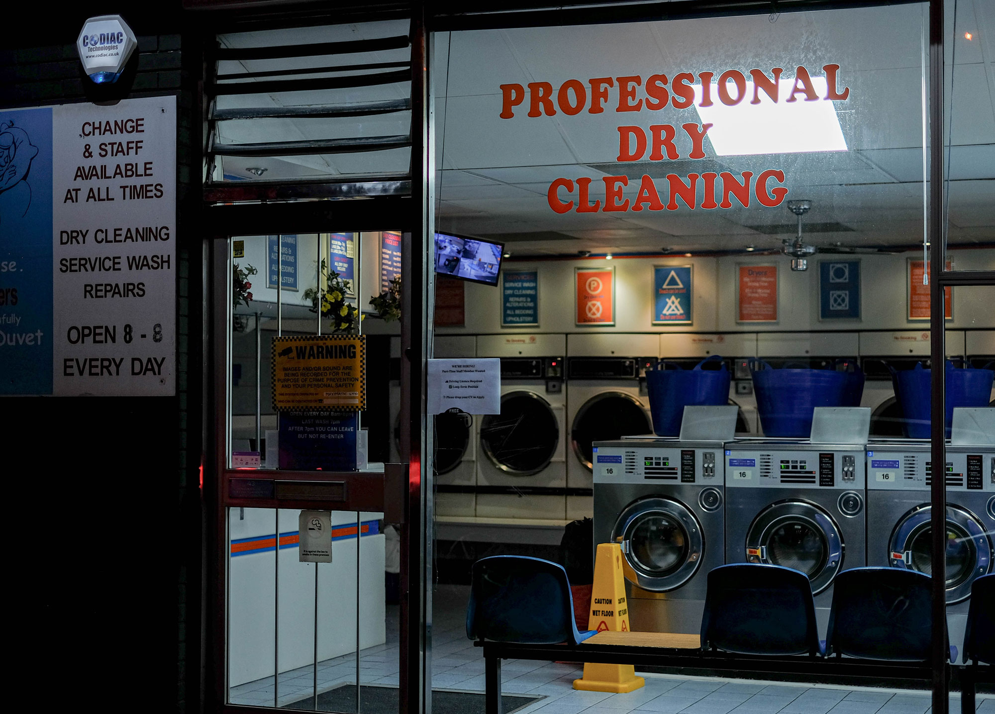 An empty laundrette seen through the window in the evening. A single yellow cone warns of a wet floor. Taken by Laura Milner.