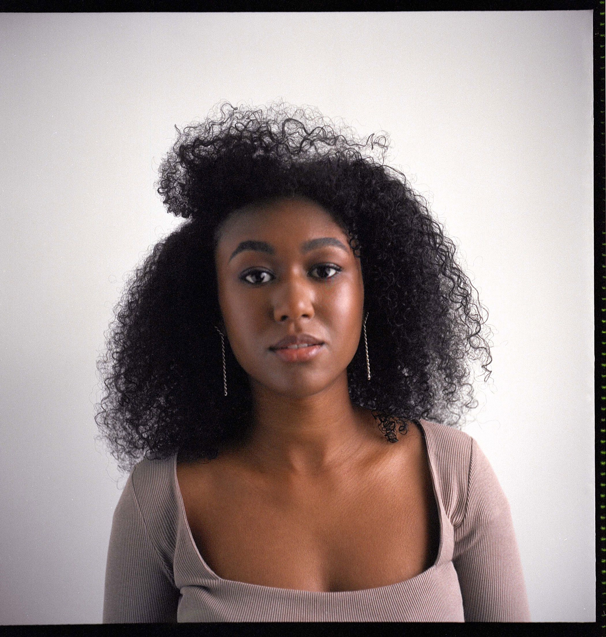 Portrait of a Black woman looking directly at the camera, against a grey background, shot by Samia Rachel.
