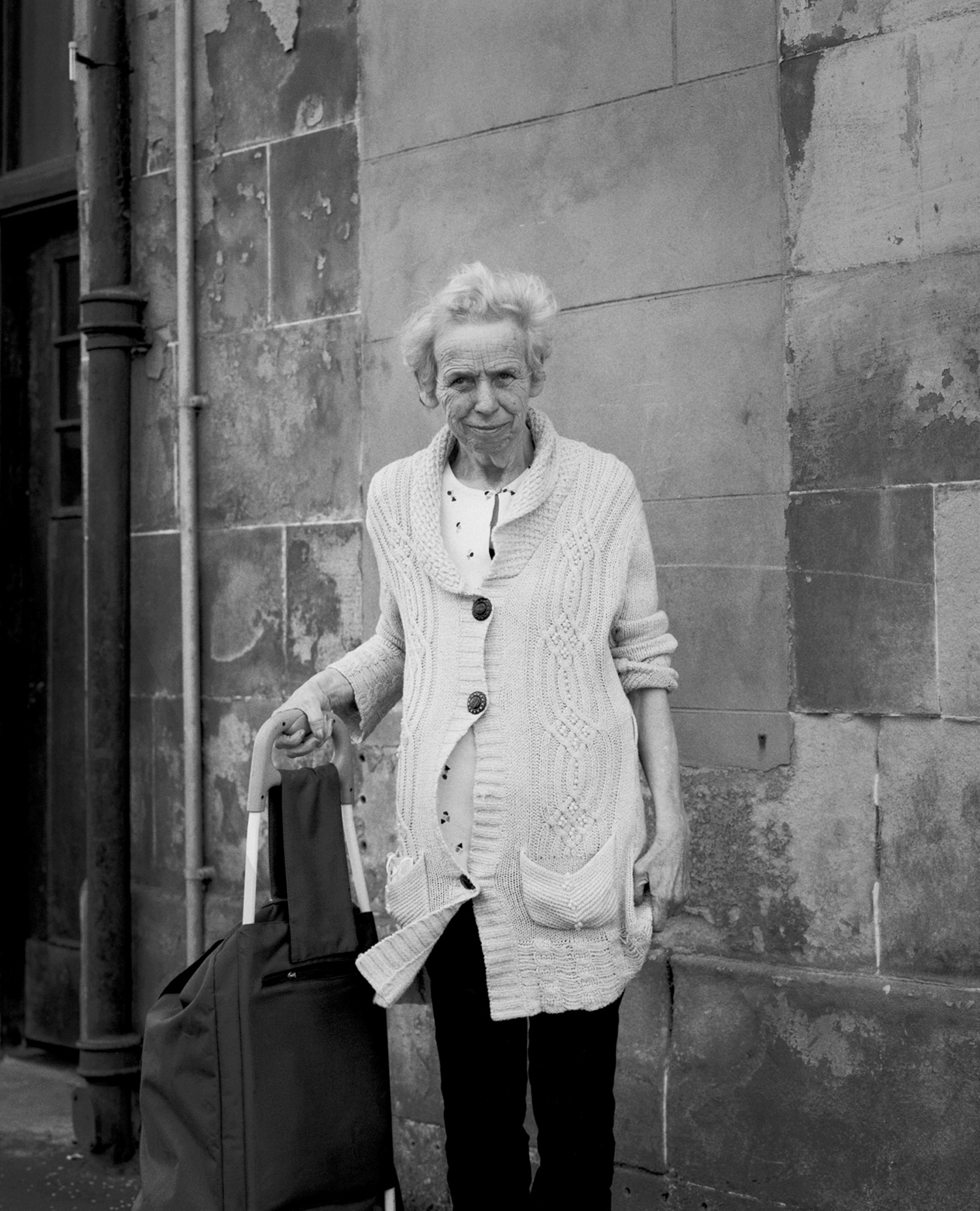 Un portrait de rue en noir et blanc d'une personne âgée avec un chariot devant un bâtiment.