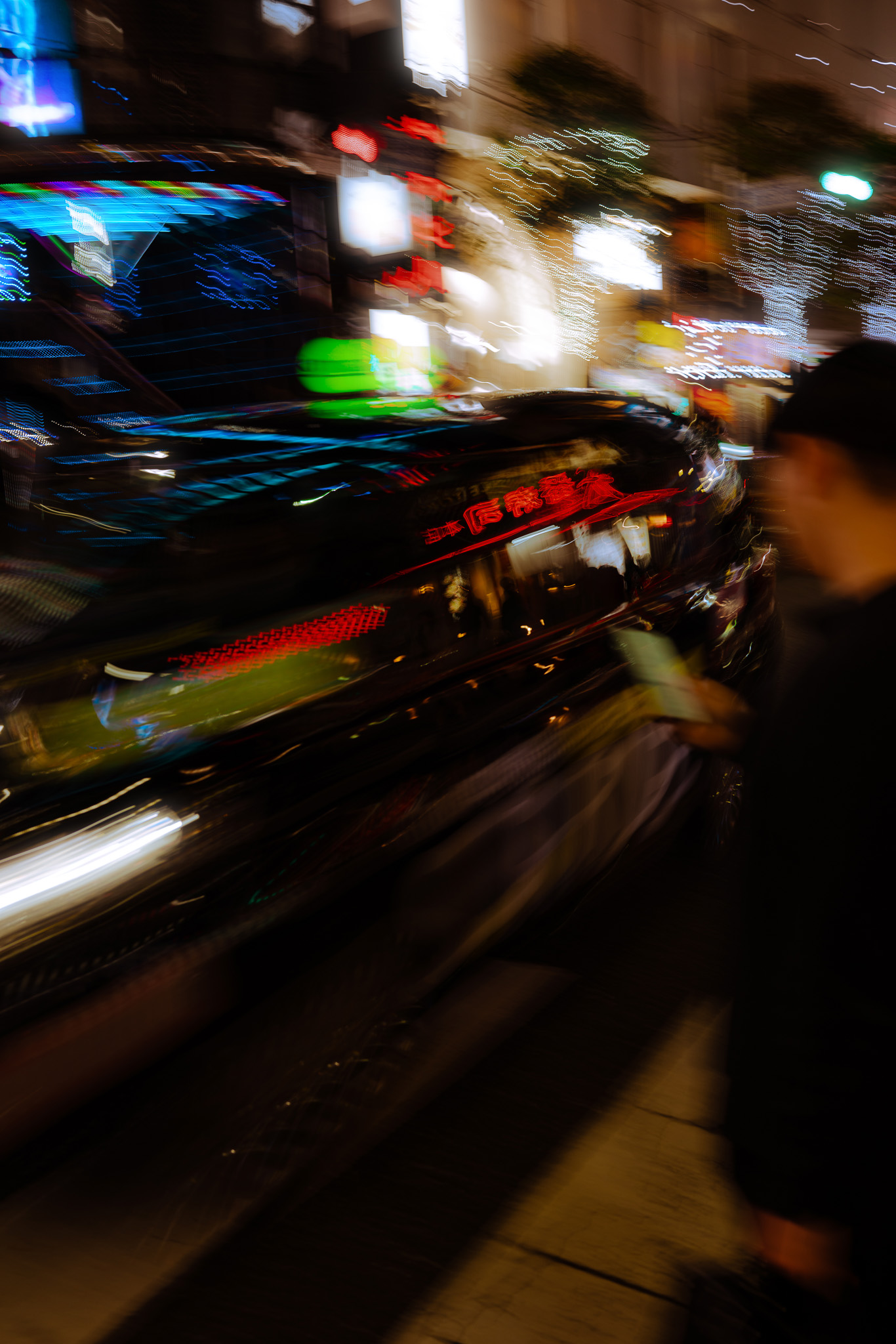 A dynamic and abstract image capturing the motion of a car reflecting vibrant neon lights in a bustling city at night, with a blurred pedestrian holding a phone. Photographed by Ruby Cruden with a Sony DSC-RX1 RM II.