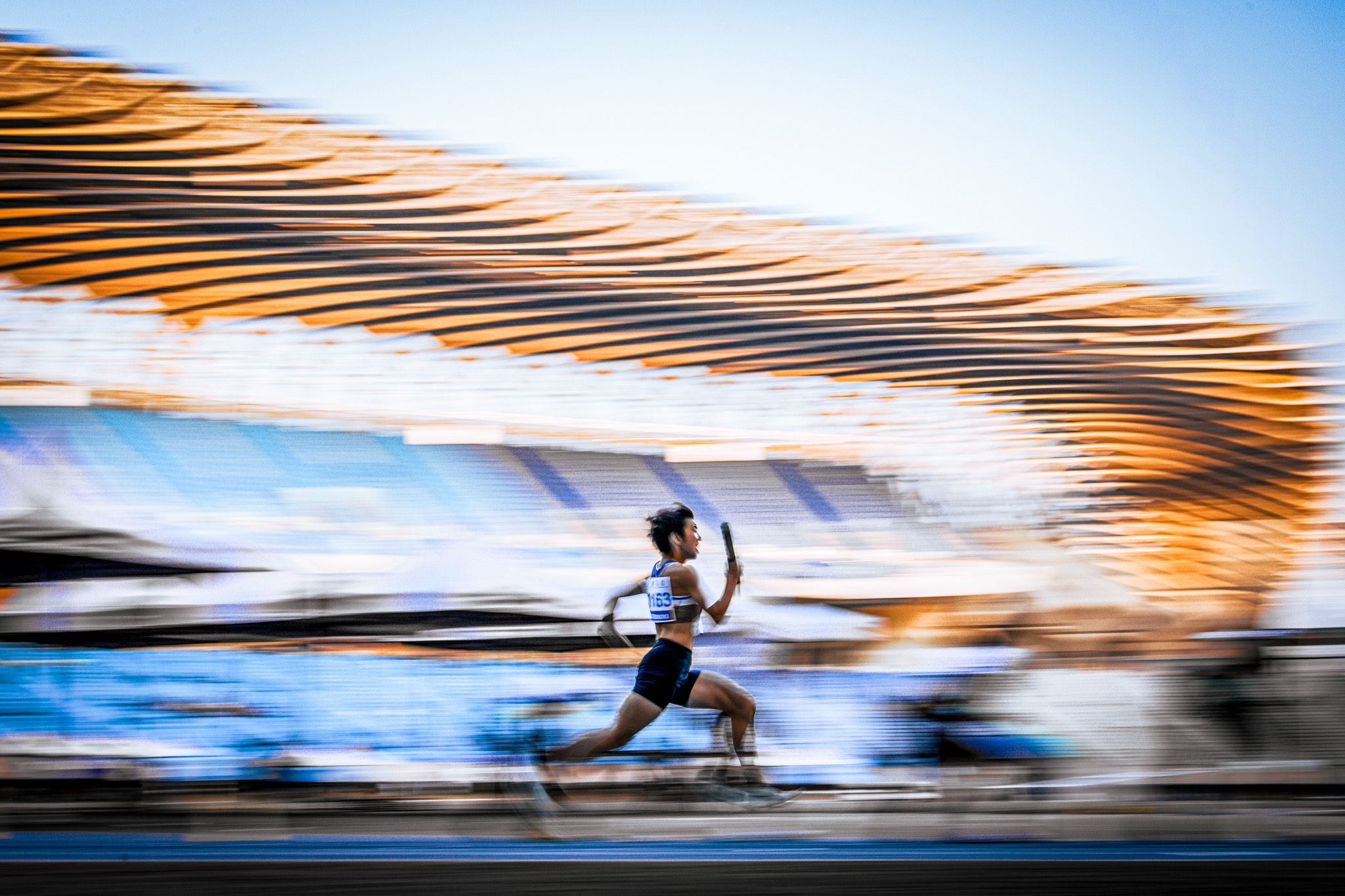 A relay runner running at full sprint with a relay baton in hand. The stadium around him is blurry. By Tetsu Lee, WSPA Gold Winner 2024. 

