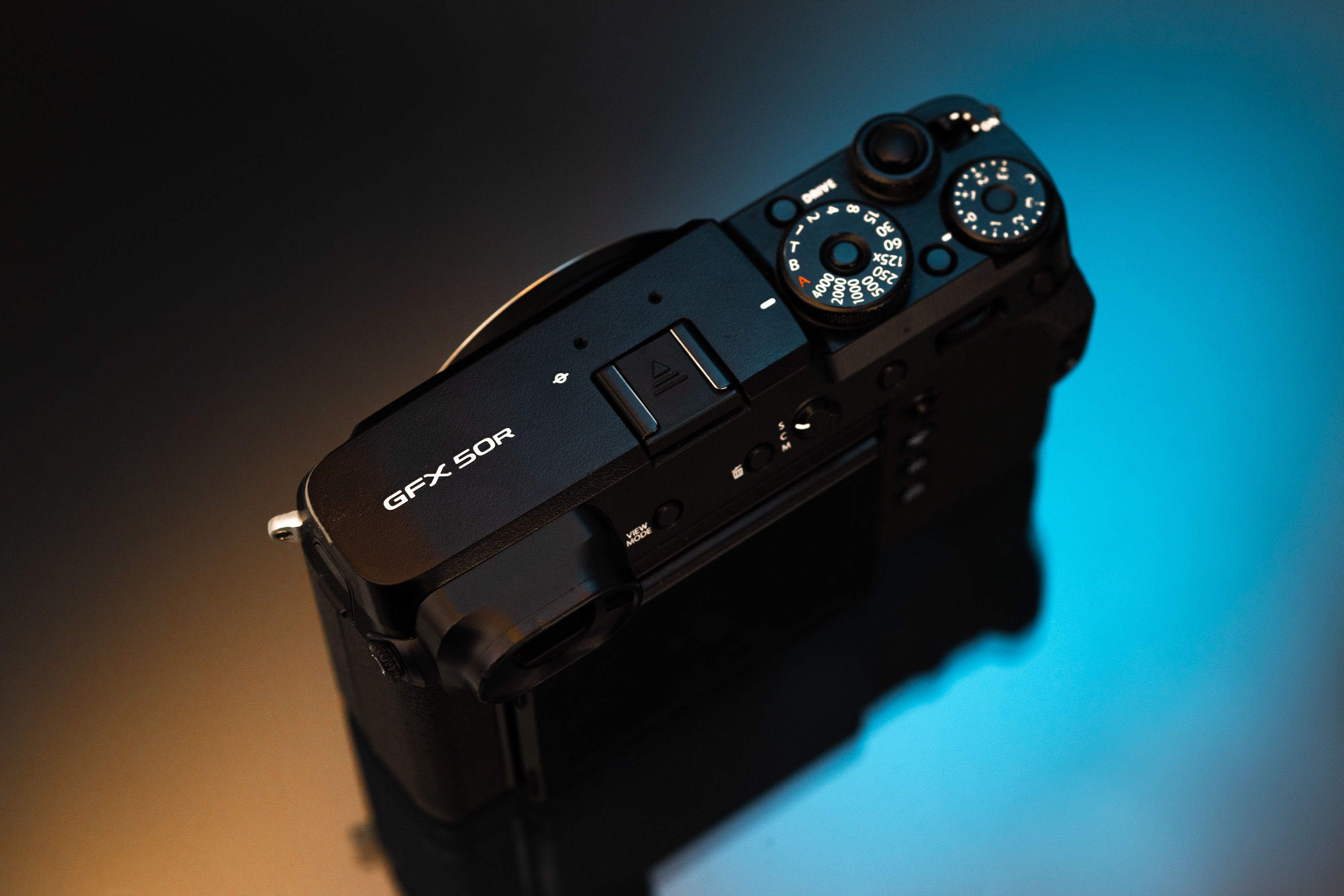 Farbfoto einer gebrauchten Fujifilm GFX 50R auf einem Glastisch, der orange und blaue Lichter reflektiert
