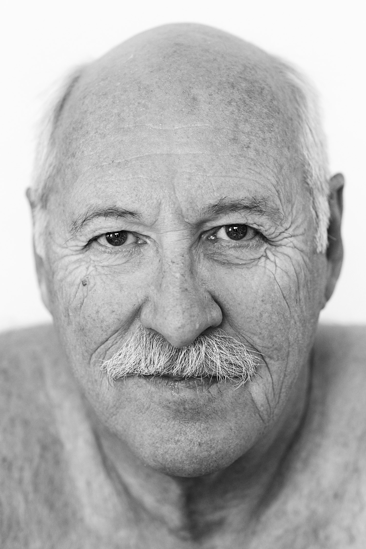 A black and white photo of a man’s face - he has a moustache and white hair. Shot by Kim Fuller.