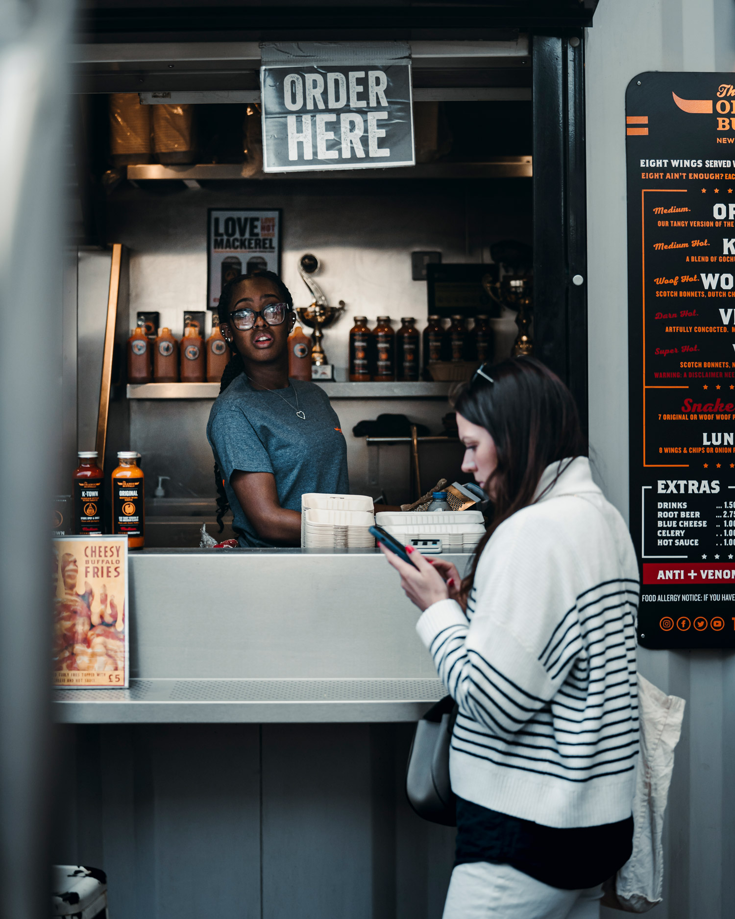 A person in a white jumper looks at their phone outside a food stall while another person stands inside, under a sign that says order here.