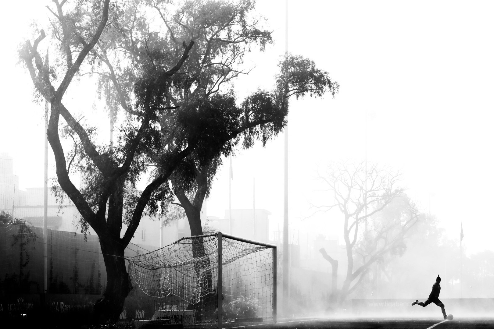 Silhouette of a football player striking a ball in the mist on a football pitch. Photo is in black and white.