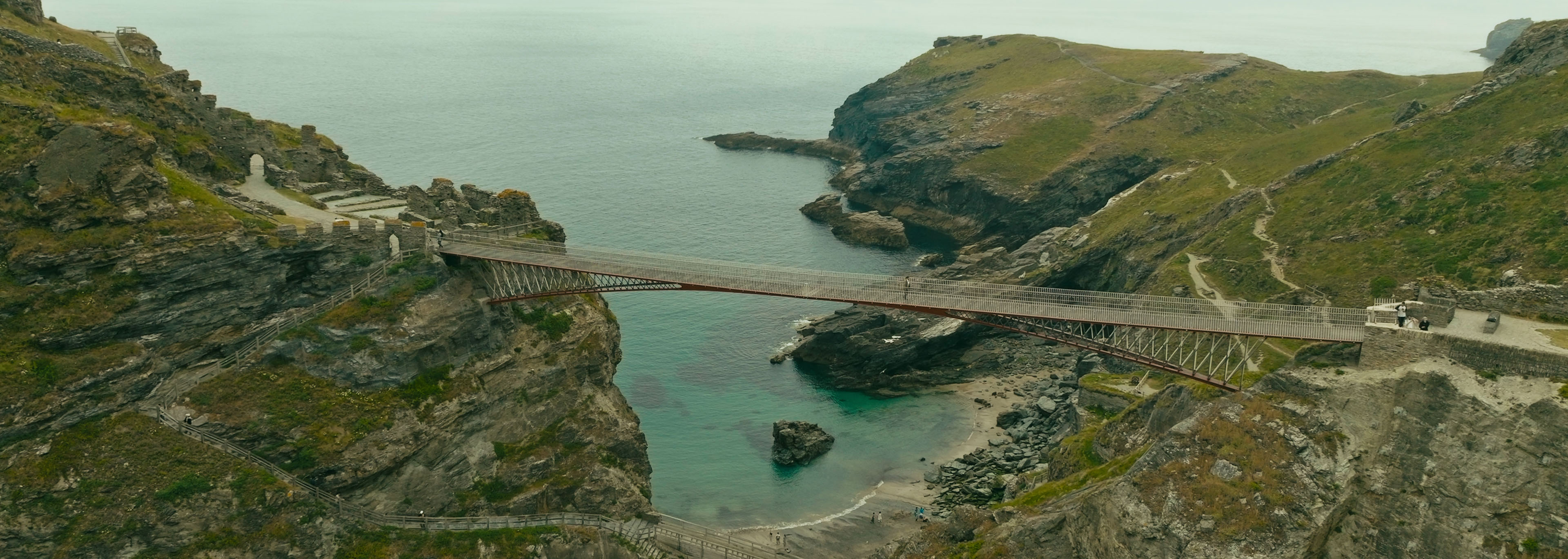 Drone aerial photo of the suspension bridge at Tintagel, Cornwall, crossing a rugged coastal inlet with cliffs and turquoise water below.