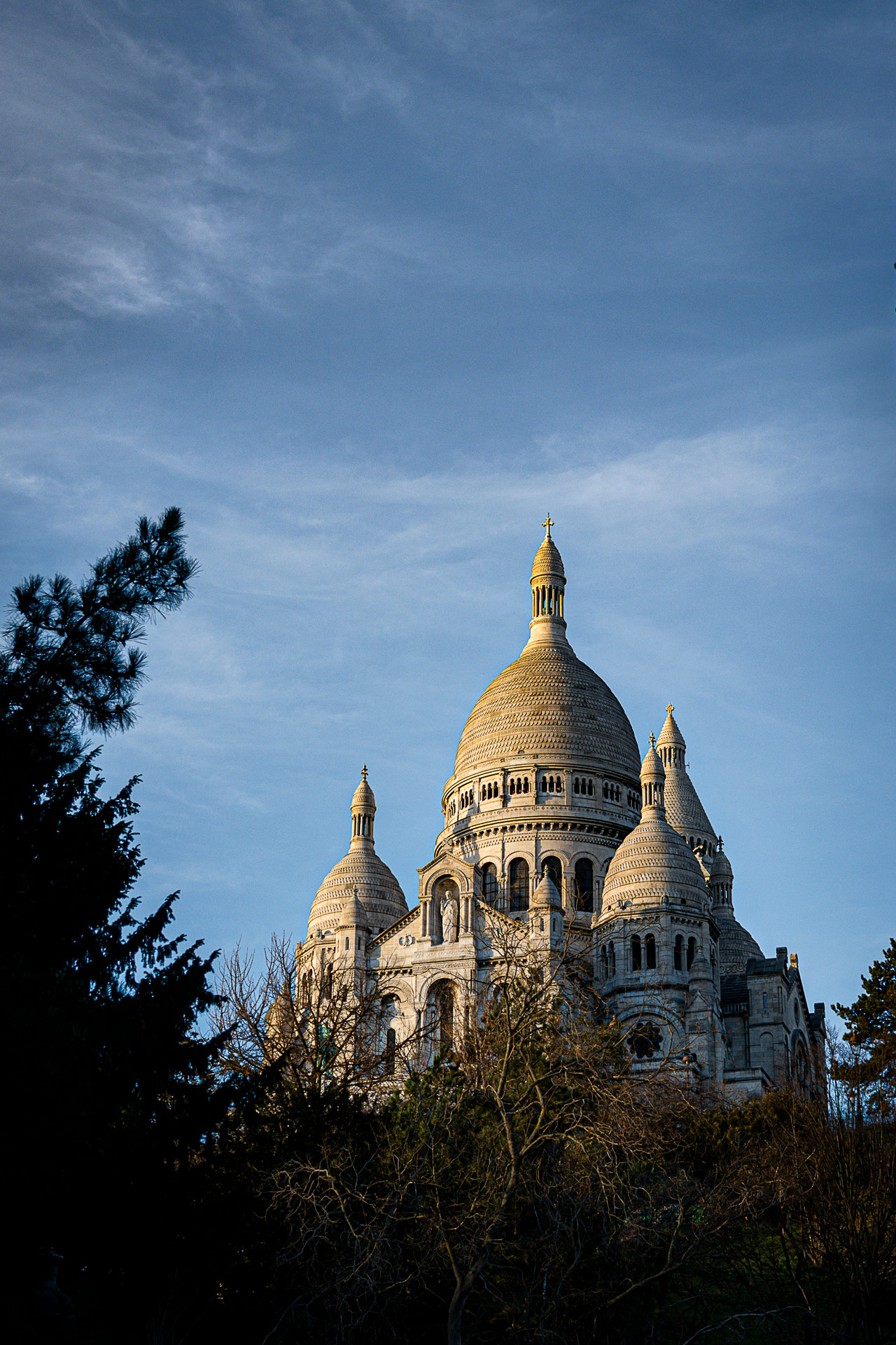 Sacré-Cœur Basilica rising above Montmartre in Paris, framed by trees under a blue sky, showcasing its white domes and historic architecture.