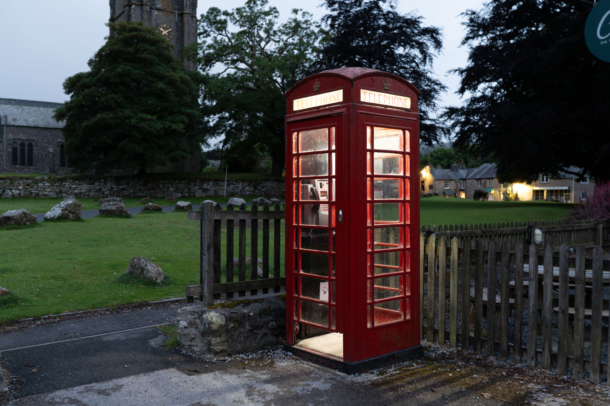 A red telephone box with a light on inside on a wet, misty day. Behind it is a fence, a church and some houses in a village. 