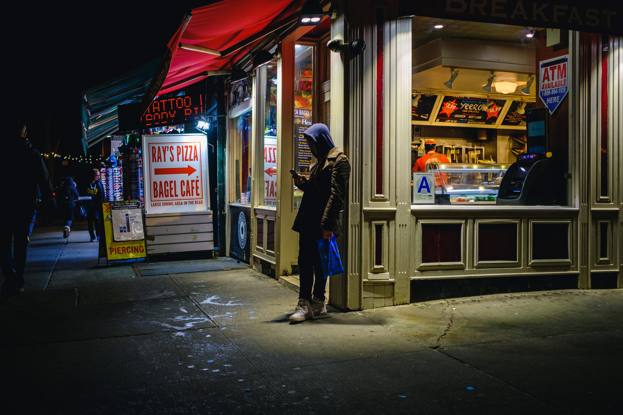 Een man leunt tegen de muur buiten een bodega in New York City en kijkt naar zijn telefoon, gefotografeerd door Manuel Pena.