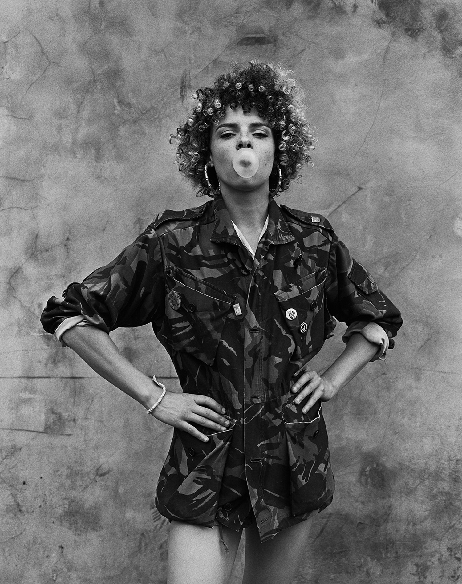 A black-and-white street portrait photo of a person with camo jacket on in front of a cracked wall, blowing a chewing gum bubble, image by Simon Murphy