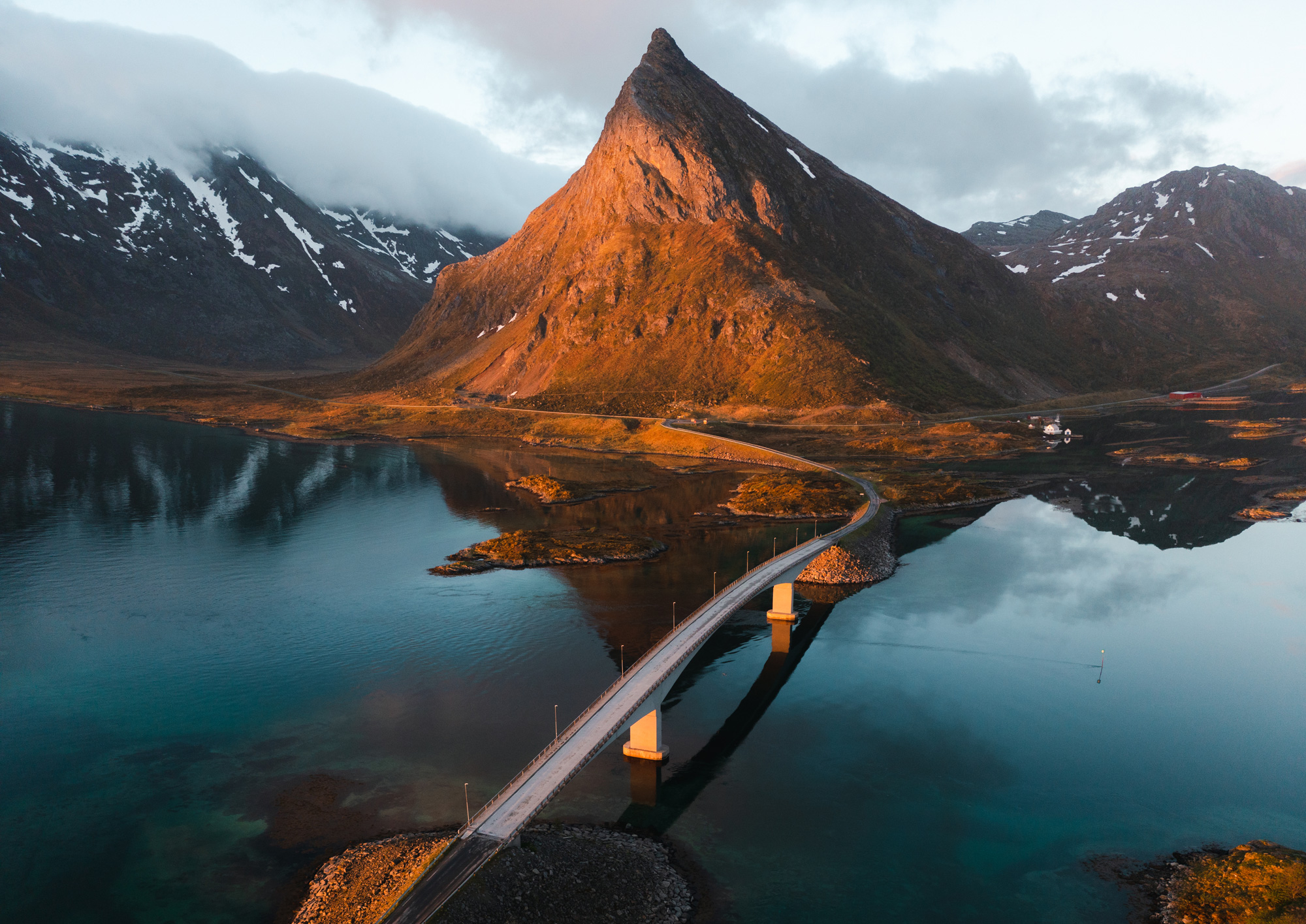 A drone photo of a bridge over water leading to a large mountain peak, surrounded by other snow-topped mountains and clouds, in Norway, shot by Jordan Lancaster-Graham with a DJI Mavic II Pro