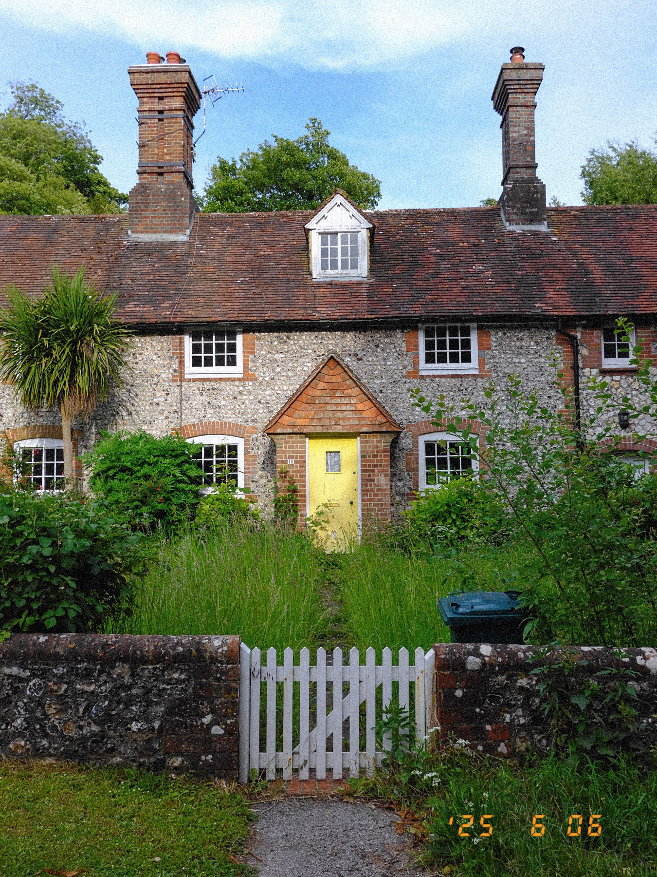 Ein Haus im Stanmer Park, Brighton, mit einer gelben Eingangstür und einem weißen Tor. Der Garten ist von langem Gras überwuchert.