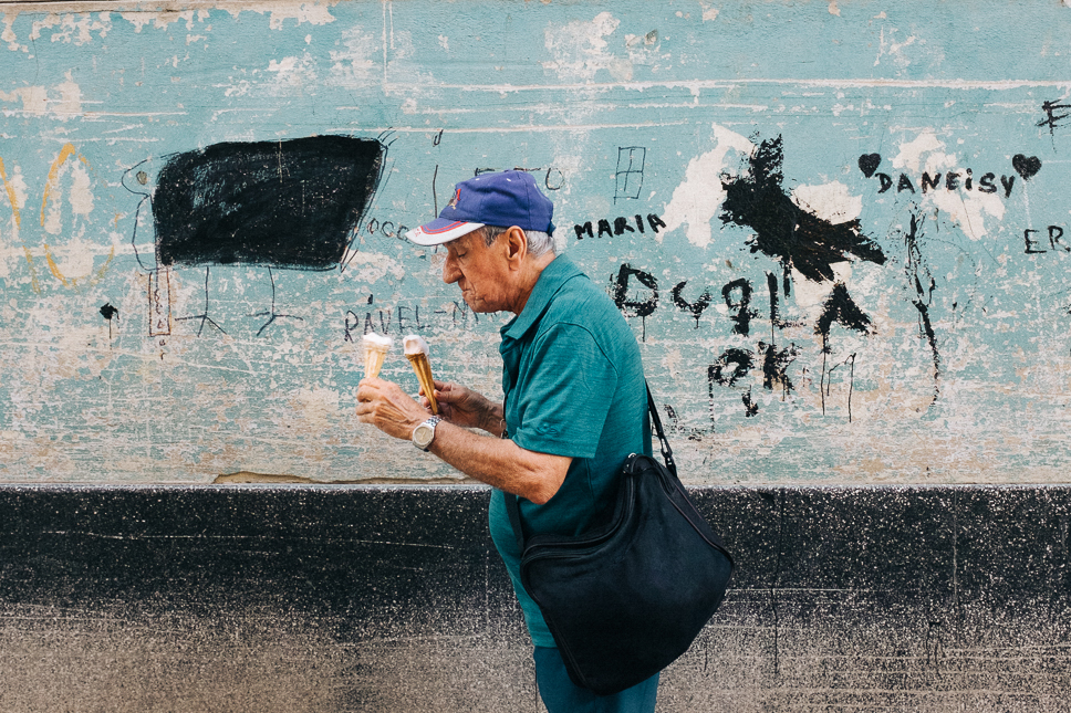 A photo of a person, wearing a turquoise polo shirt, blue hat and black bag, eating two ice creams in front of a light-blue wall, covered in black graffiti, street photography by Ian Howorth