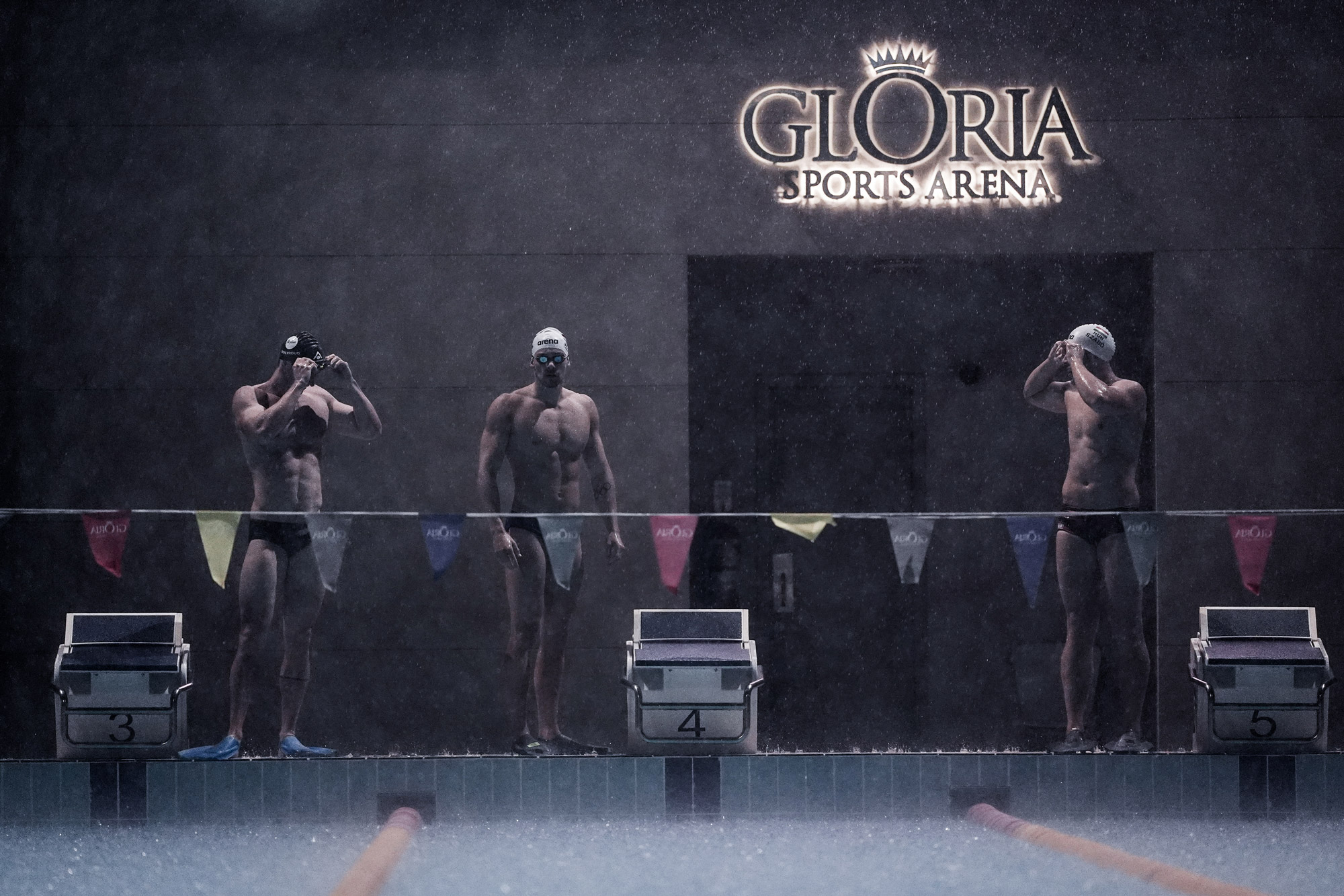 Three swimmers stand in preparation in front of a swimming pool with race markers and behind a string of bunting.
