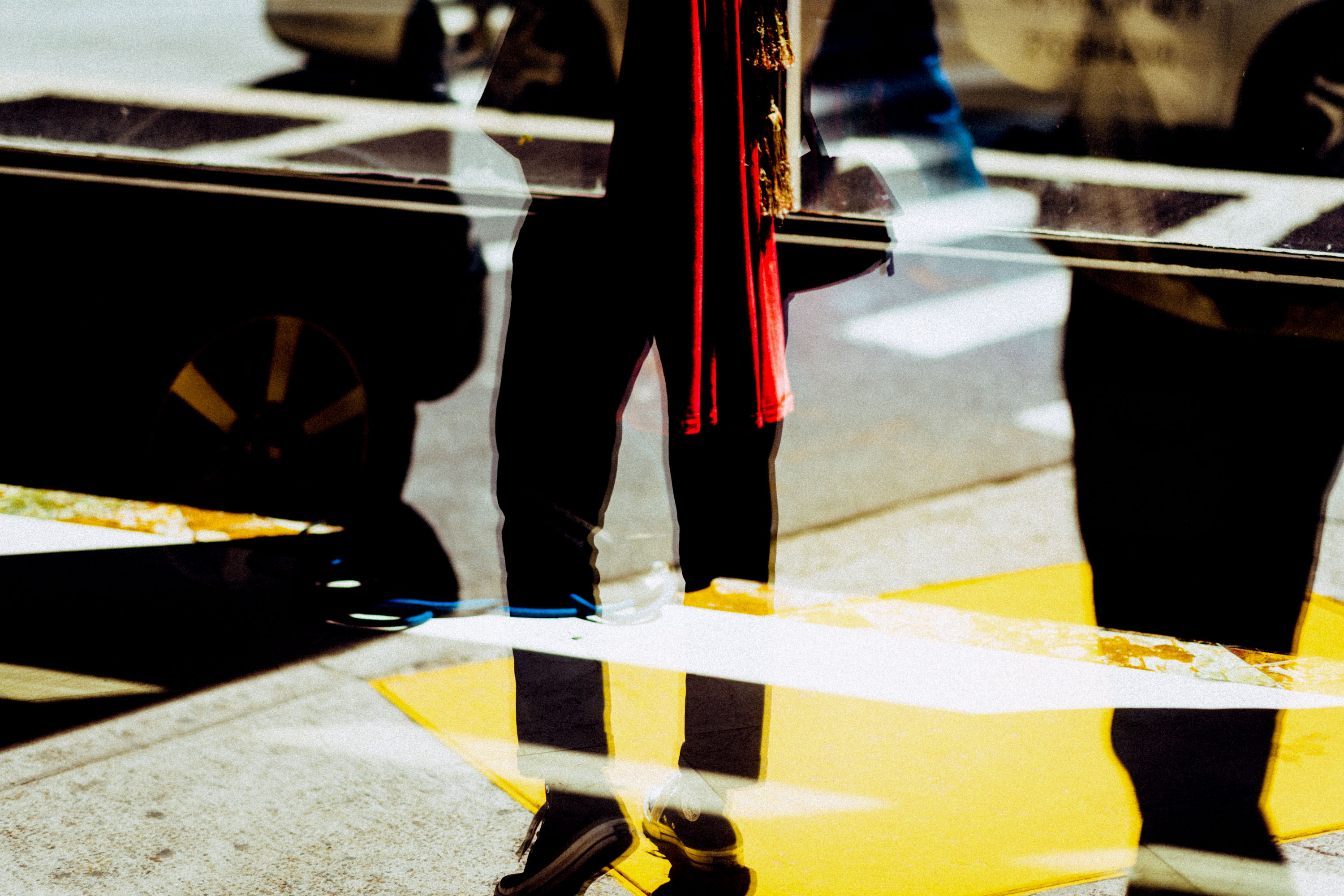 A mirrored image of a pair of legs in front of a pavement with a flash of yellow in the frame, street photography by Ian Howorth
