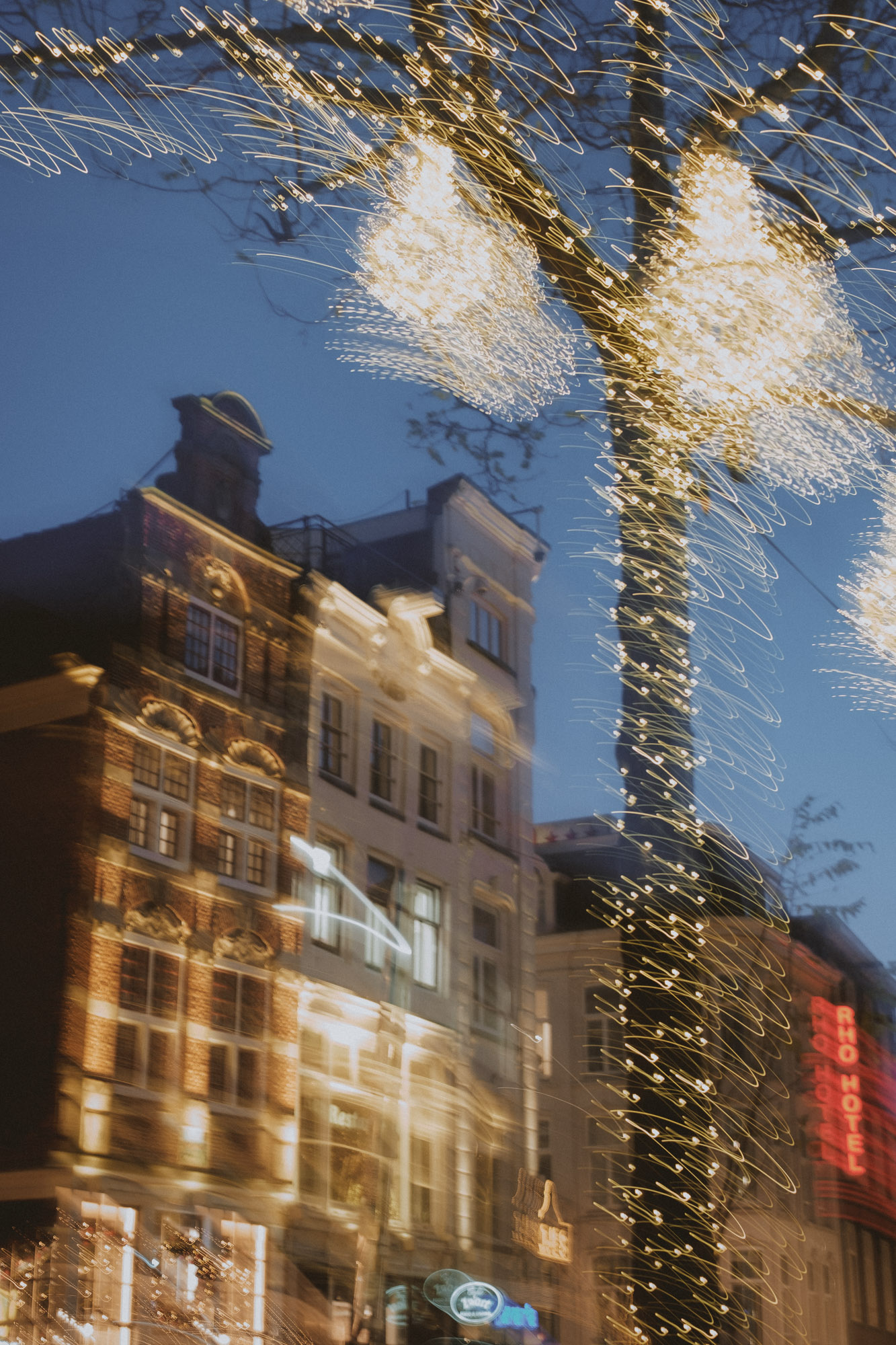Maisons anciennes bordant les canaux d'Amsterdam, illuminées à la tombée de la nuit par des guirlandes festives enroulées autour des arbres, photographiées avec un effet de flou de mouvement créant une scène urbaine chaleureuse et pleine d'ambiance