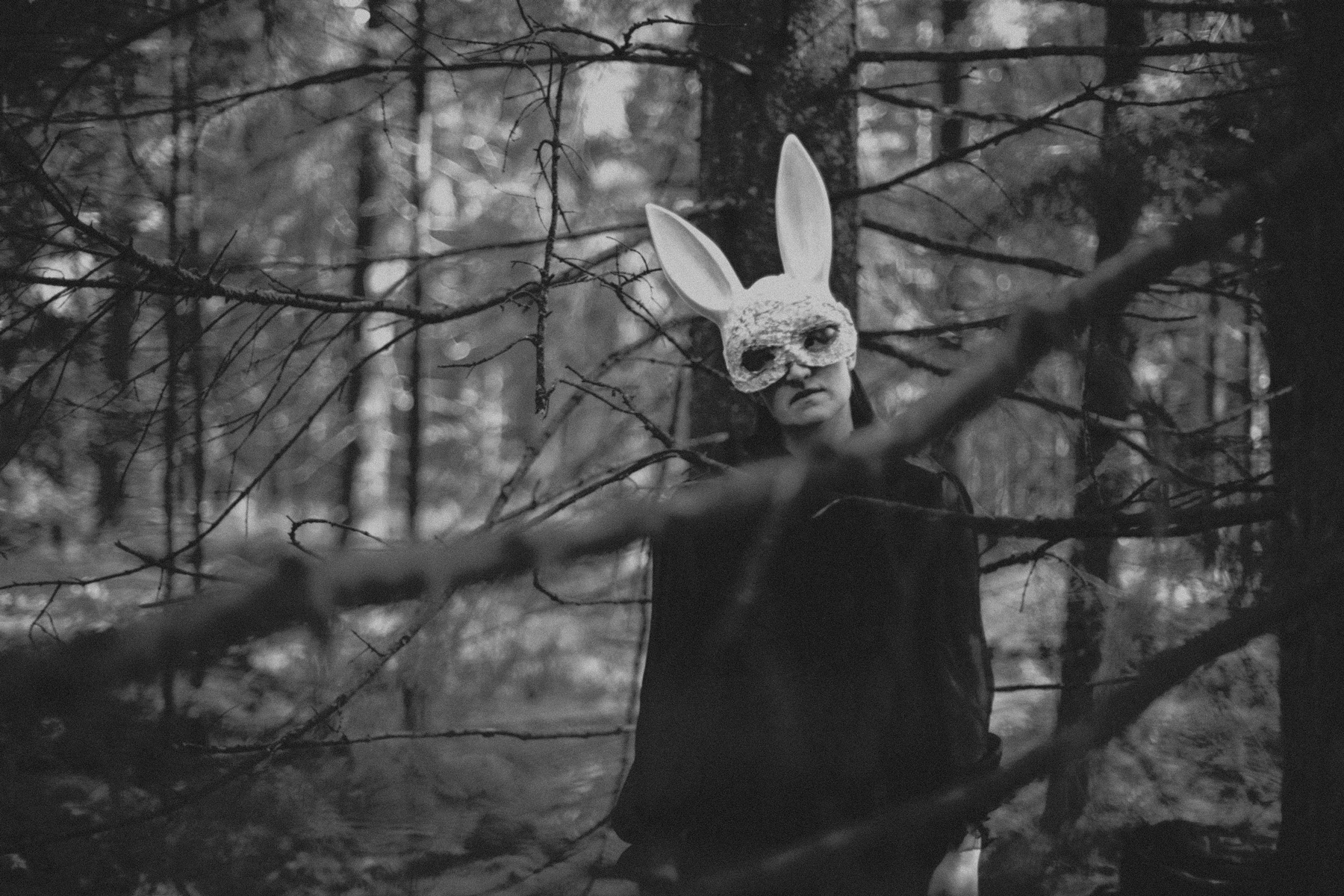 Ein Schwarz-Weiß-Foto eines Modells in einem dunklen Hemd und einer Hasenmaske, das im Wald steht und durch Kiefernzweige in die Kamera blickt.