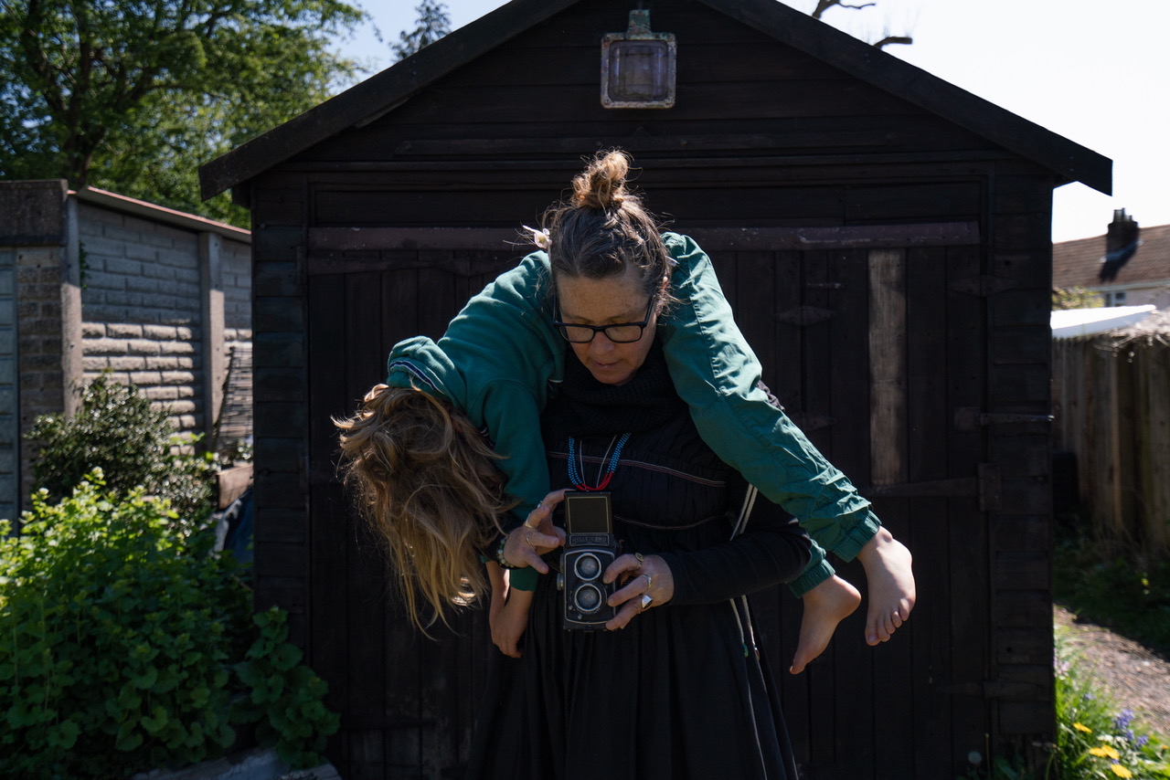 Karni Arieli holding their child over their shoulders while taking a photo with a medium-format camera outdoors in front of a shed