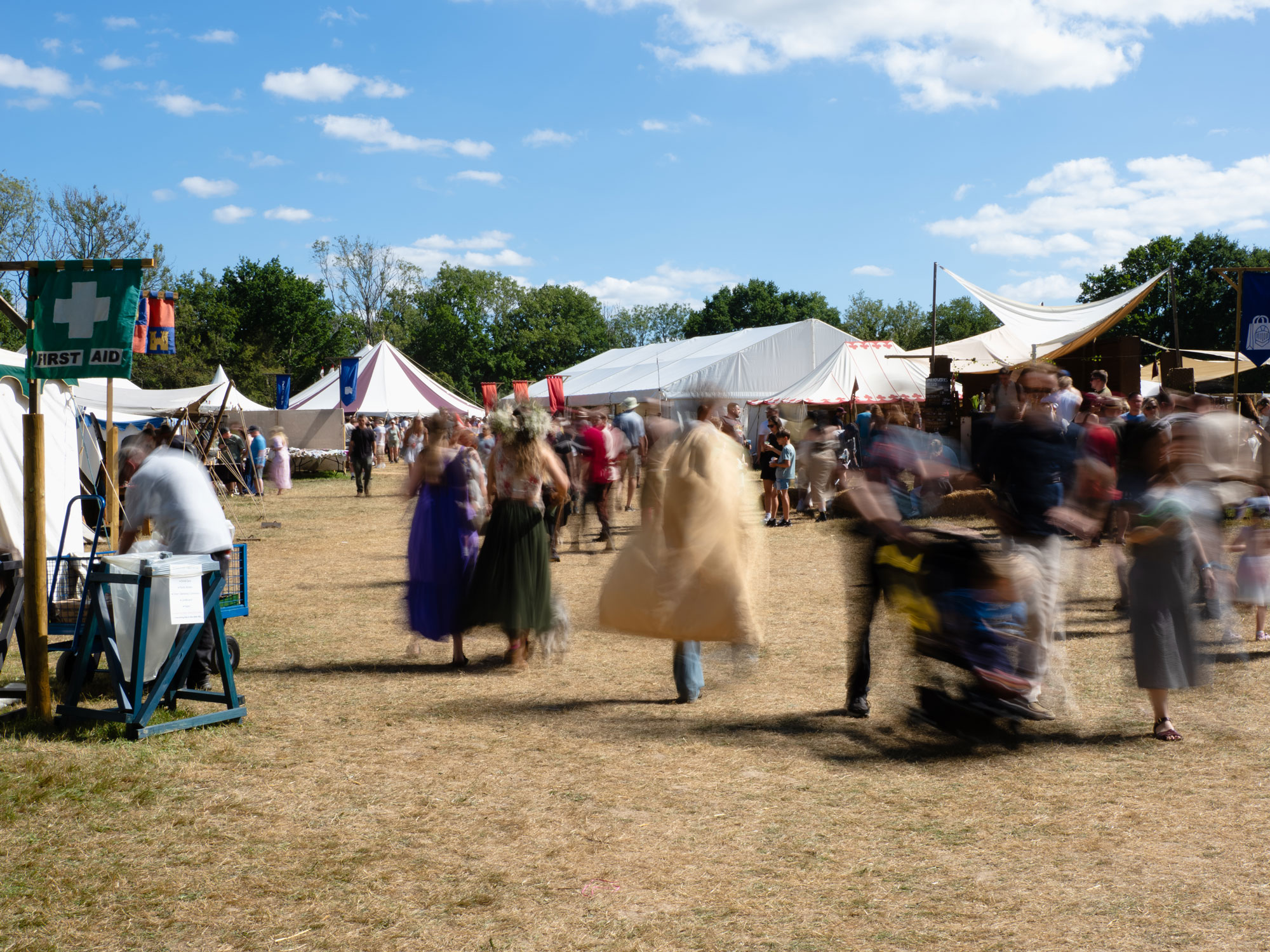 A crowd at a medieval festival, with bell tents in the background. It is a sunny day, and the crowd are dressed in costume, with those in the foreground blurred due to motion.