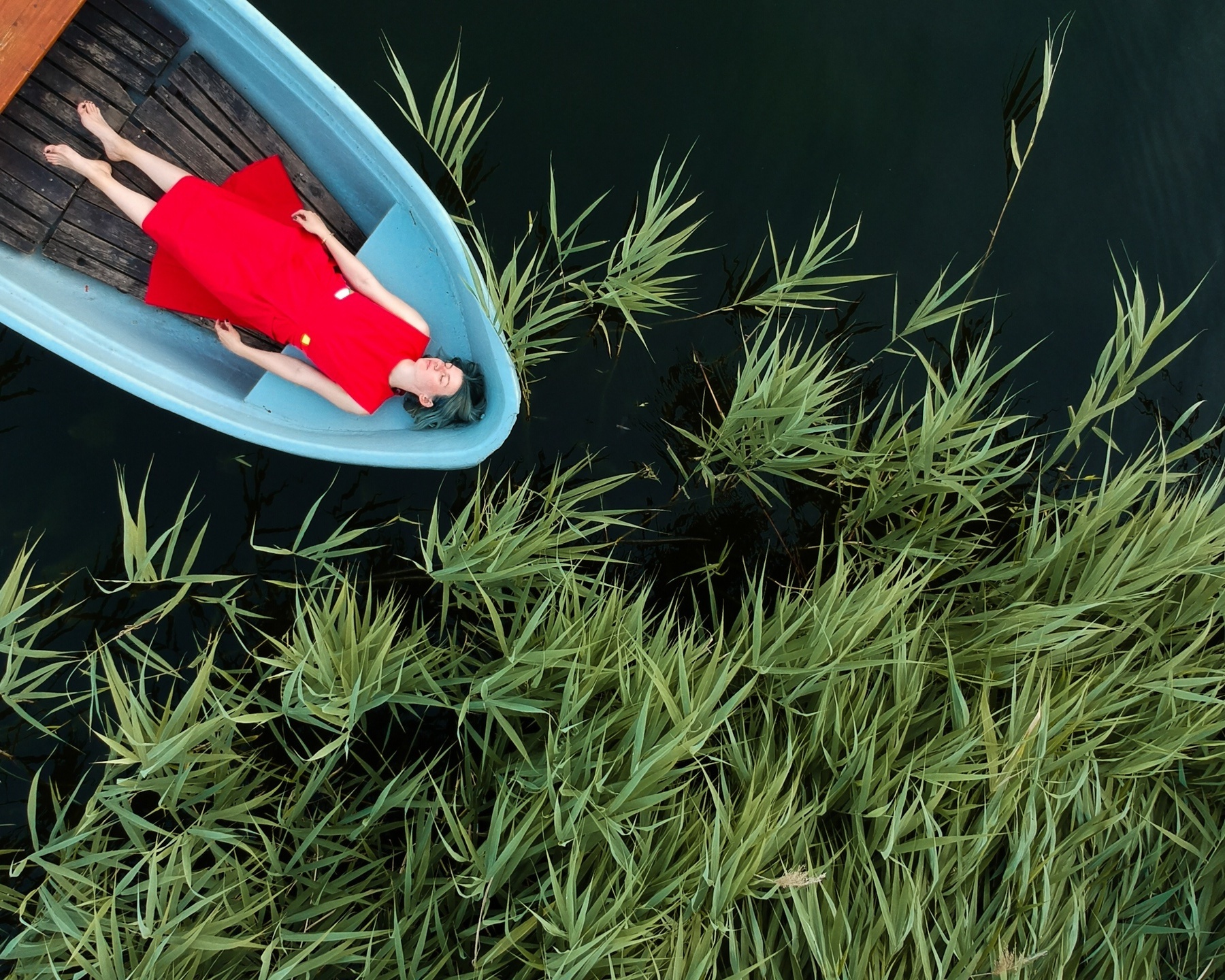 A person in a bright red dress lying peacefully in a small blue rowboat on dark water, surrounded by tall green reeds. Shot by Alina Rudya on a DJI FC220.