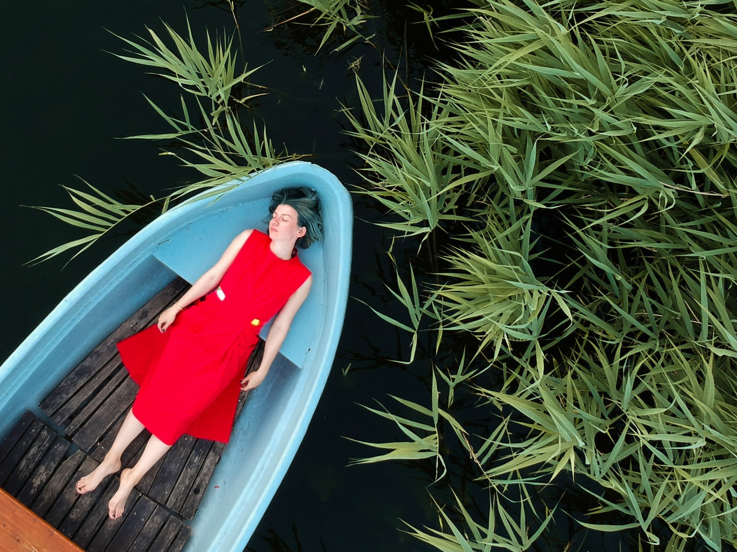 A person in a bright red dress lying peacefully in a small blue rowboat on dark water, surrounded by tall green reeds. Shot by Alina Rudya on a DJI FC220.