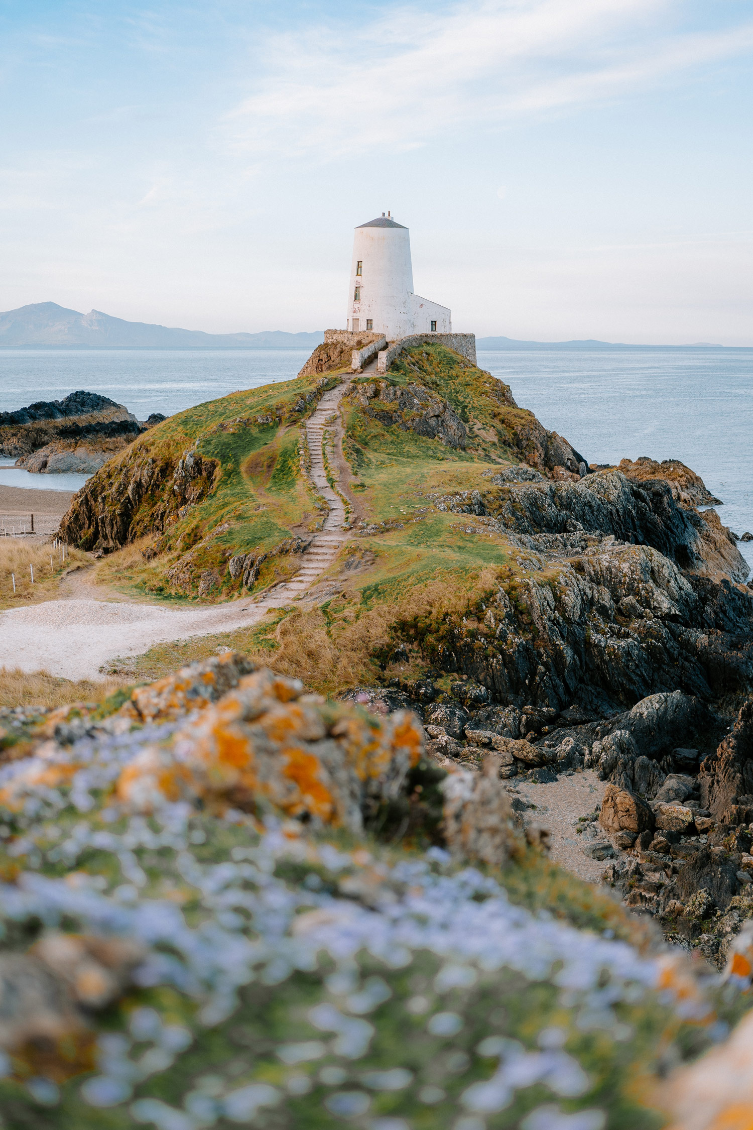 A path leads up to a lighthouse with grass and flowers in the foreground at Newborough Beach in North Wales, shot with the Sony A7C by James Popsys

