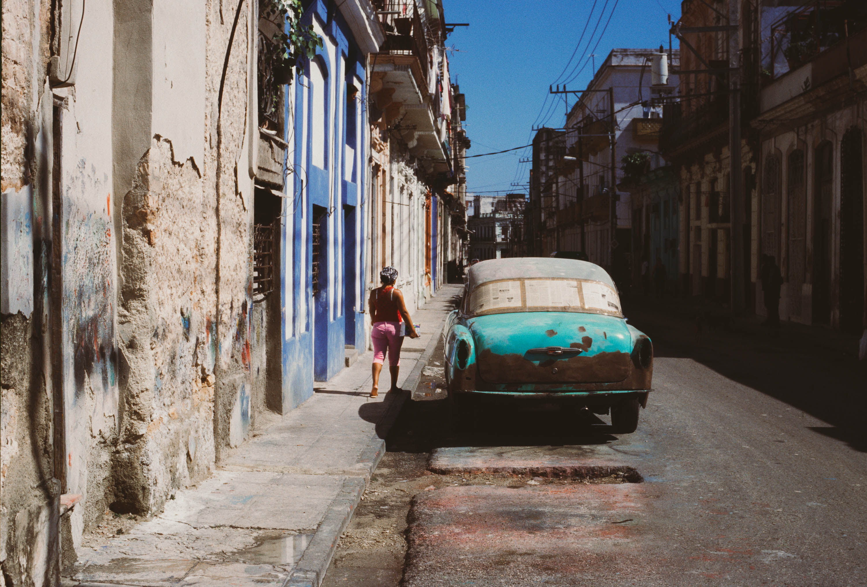 A person wearing pink walks away on a pavement with a rusty turquoise parked car and colourful buildings on either side of the road, street photography by Ian Howorth