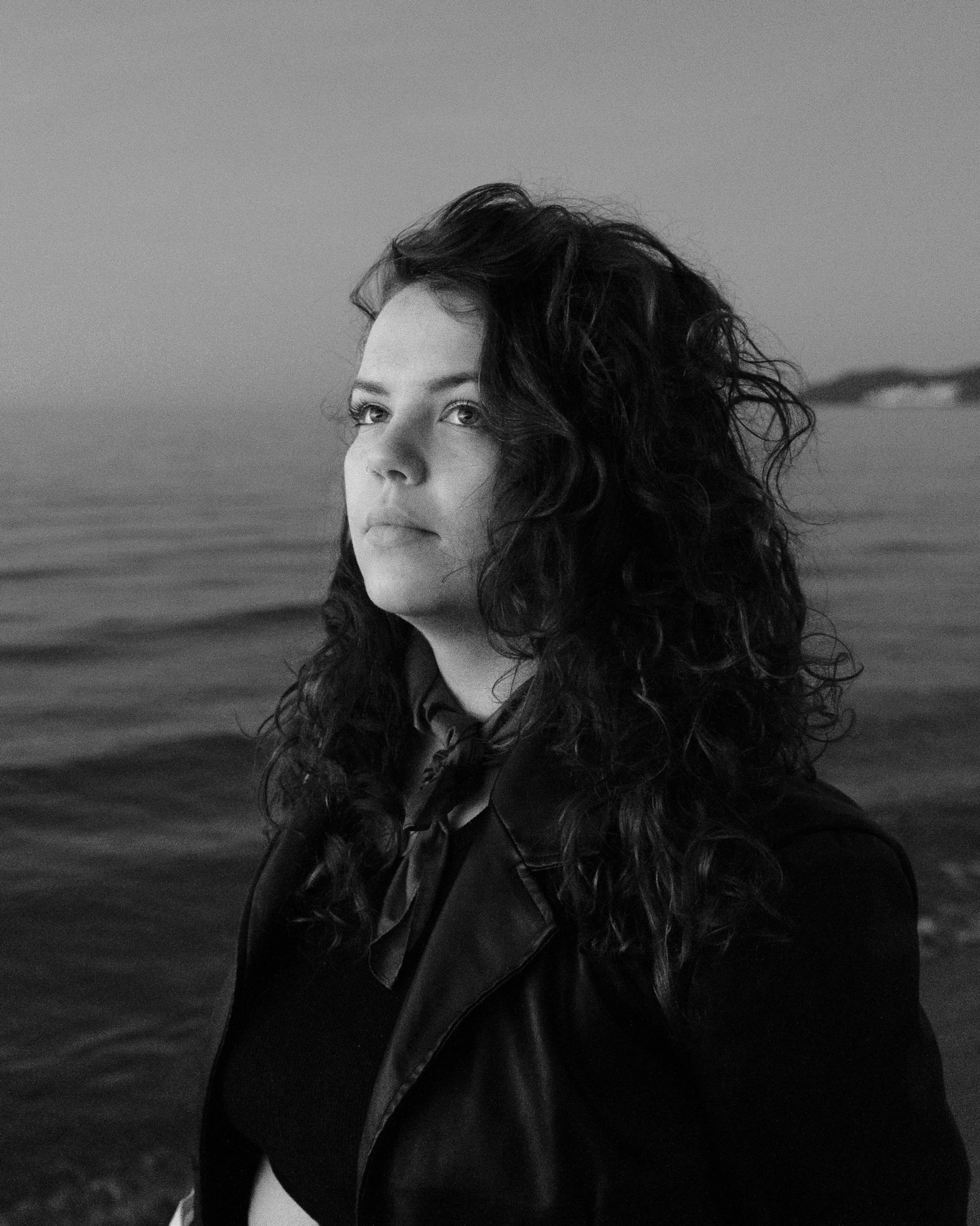 
Schwarz-Weiß-Porträt einer Person mit dunklen Haaren und dunkler Kleidung, die an der Kamera vorbeischaut, im Hintergrund Meer. 

