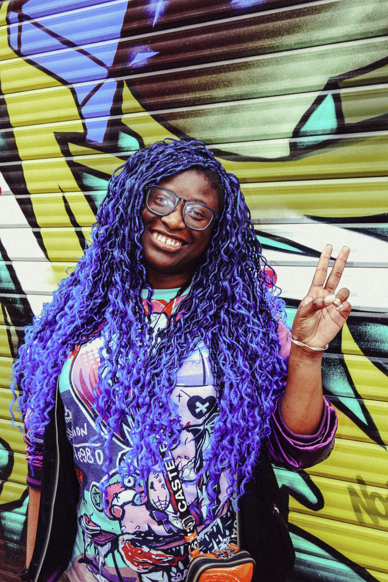 Streetporträt einer lächelnden Person mit langen lila Haaren, die vor einem bunten, mit Graffiti bedeckten Metallrollladen in einer Pariser Straße posiert und ein Peace-Zeichen macht. Aufgenommen von Wesley Verhoeve mit einer Panasonic Lumix GF1.