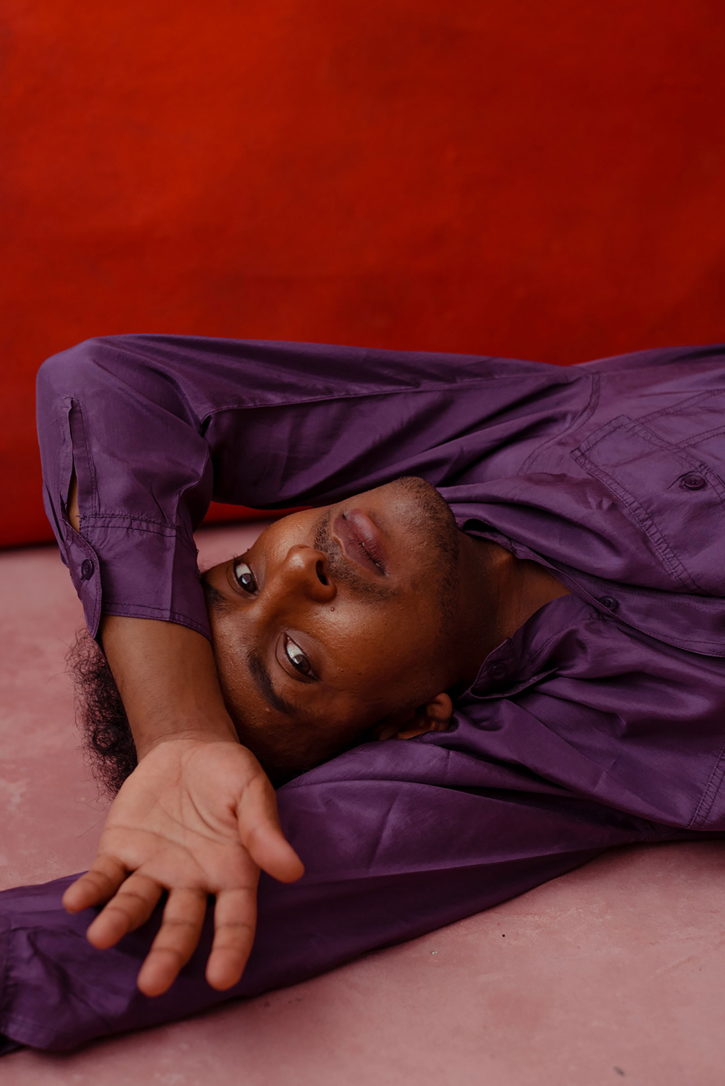 Black man lying on the floor with his left arm resting on his forehead in front of a red background, shot by Nicole Benewaah.
